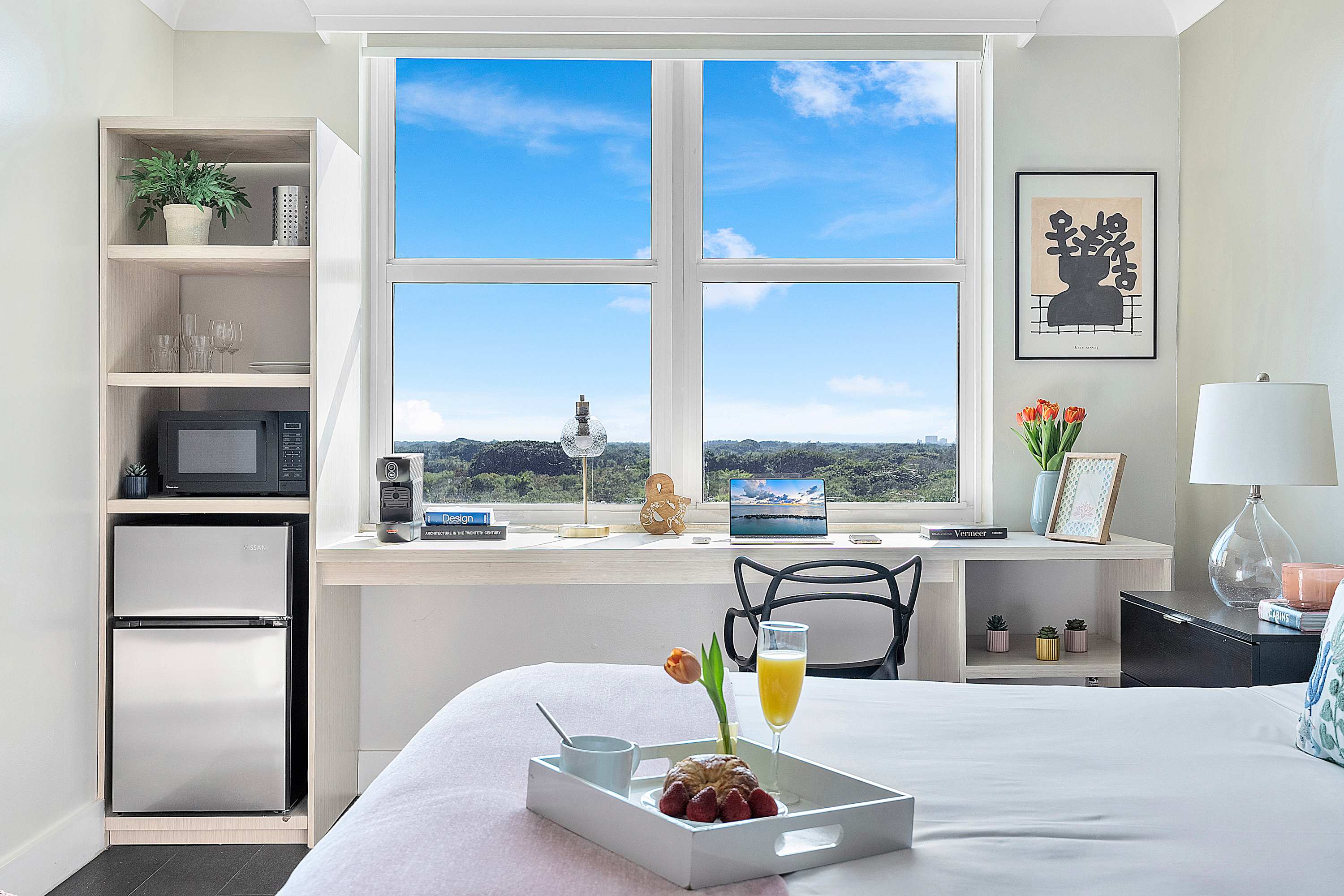 Bright room featuring a bed positioned against a large window with a green landscape view, a built-in desk with a chair, and a shelving unit housing a microwave and mini-fridge.