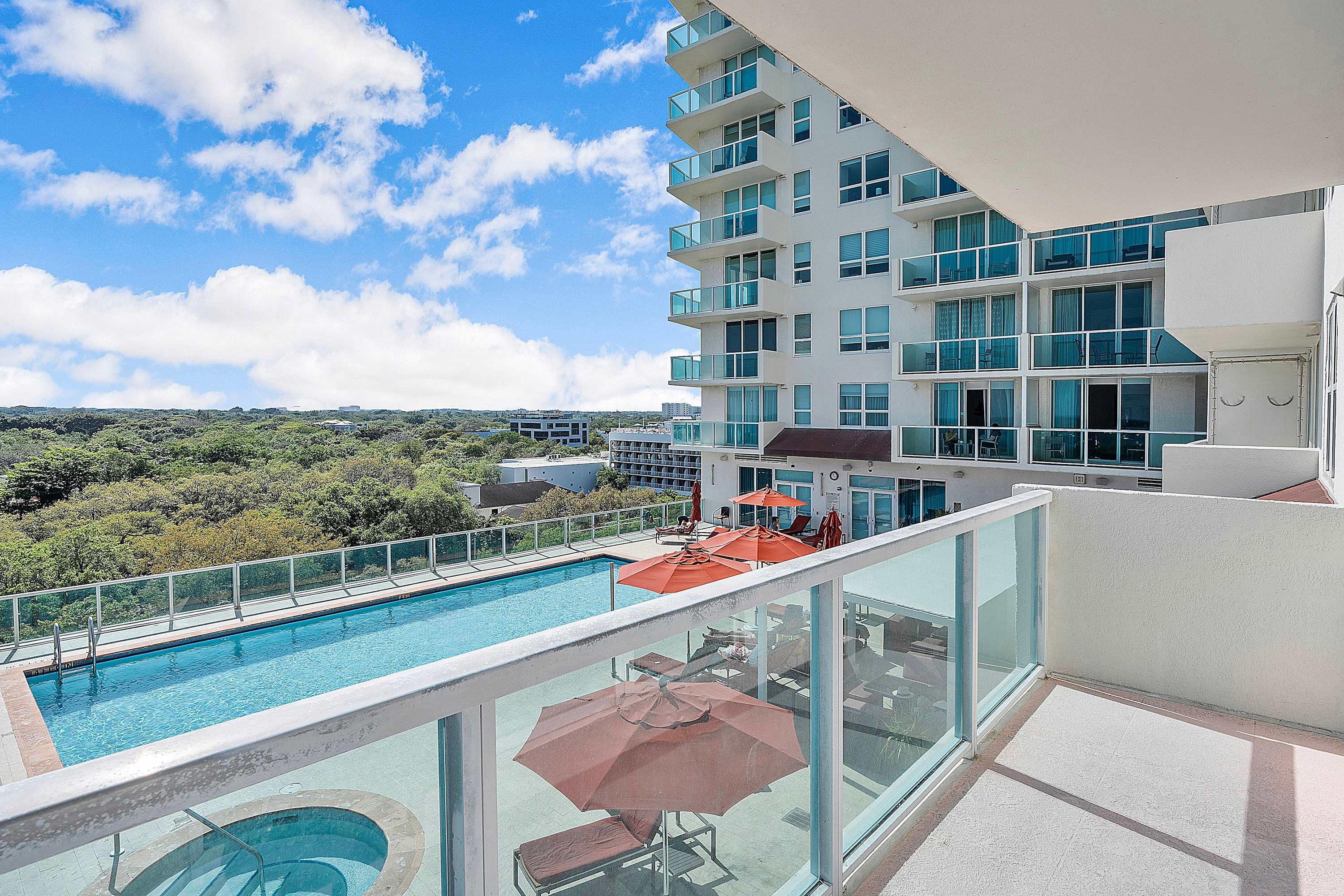 Balcony view overlooking a modern high-rise building and a resort-style pool area featuring a rectangular swimming pool, a circular hot tub, and several lounge chairs with orange umbrellas.