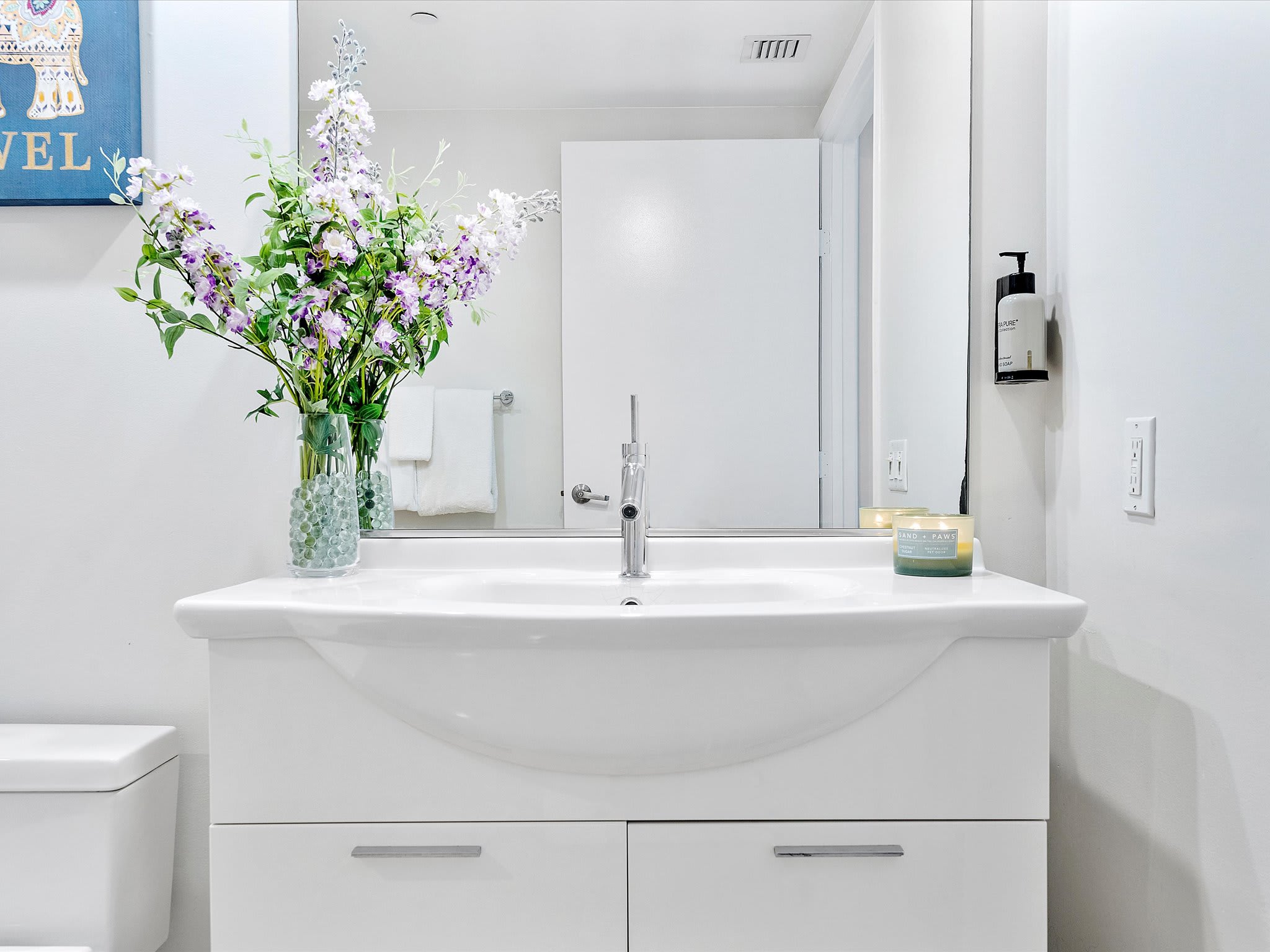 Bright bathroom featuring a white single vanity with an integrated sink and a modern chrome faucet. A large mirror hangs above, reflecting a white door, and a vase of purple and white flowers adds a pop of color.