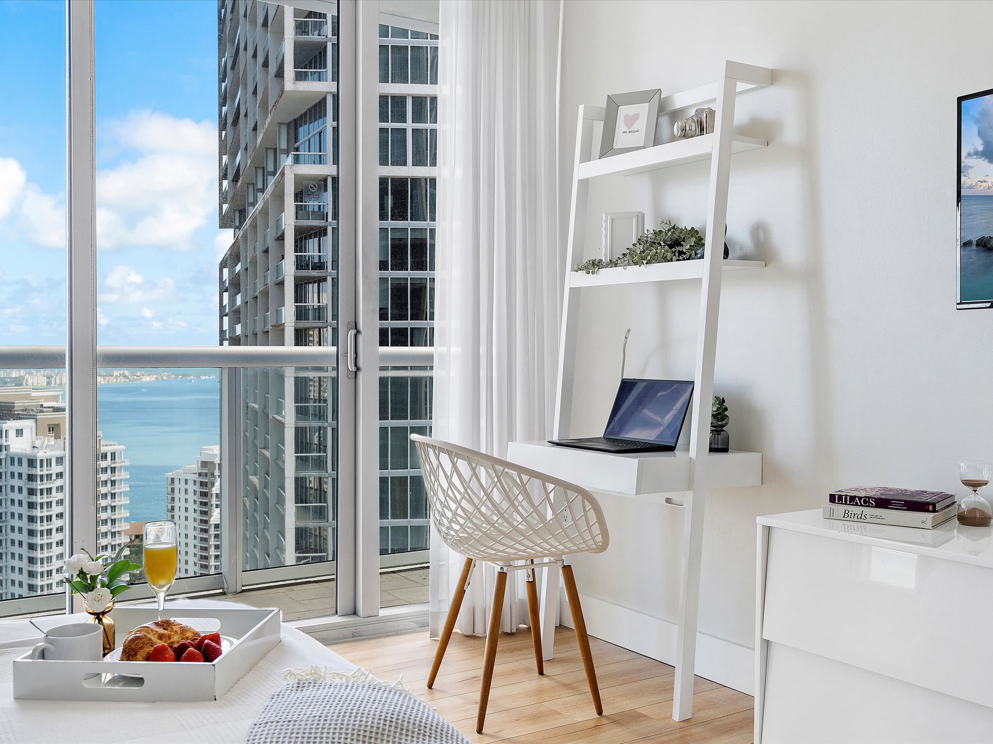 A dedicated workspace within the second bedroom, featuring a white ladder desk with a laptop, a modern chair, and large windows providing views of the city and bay.