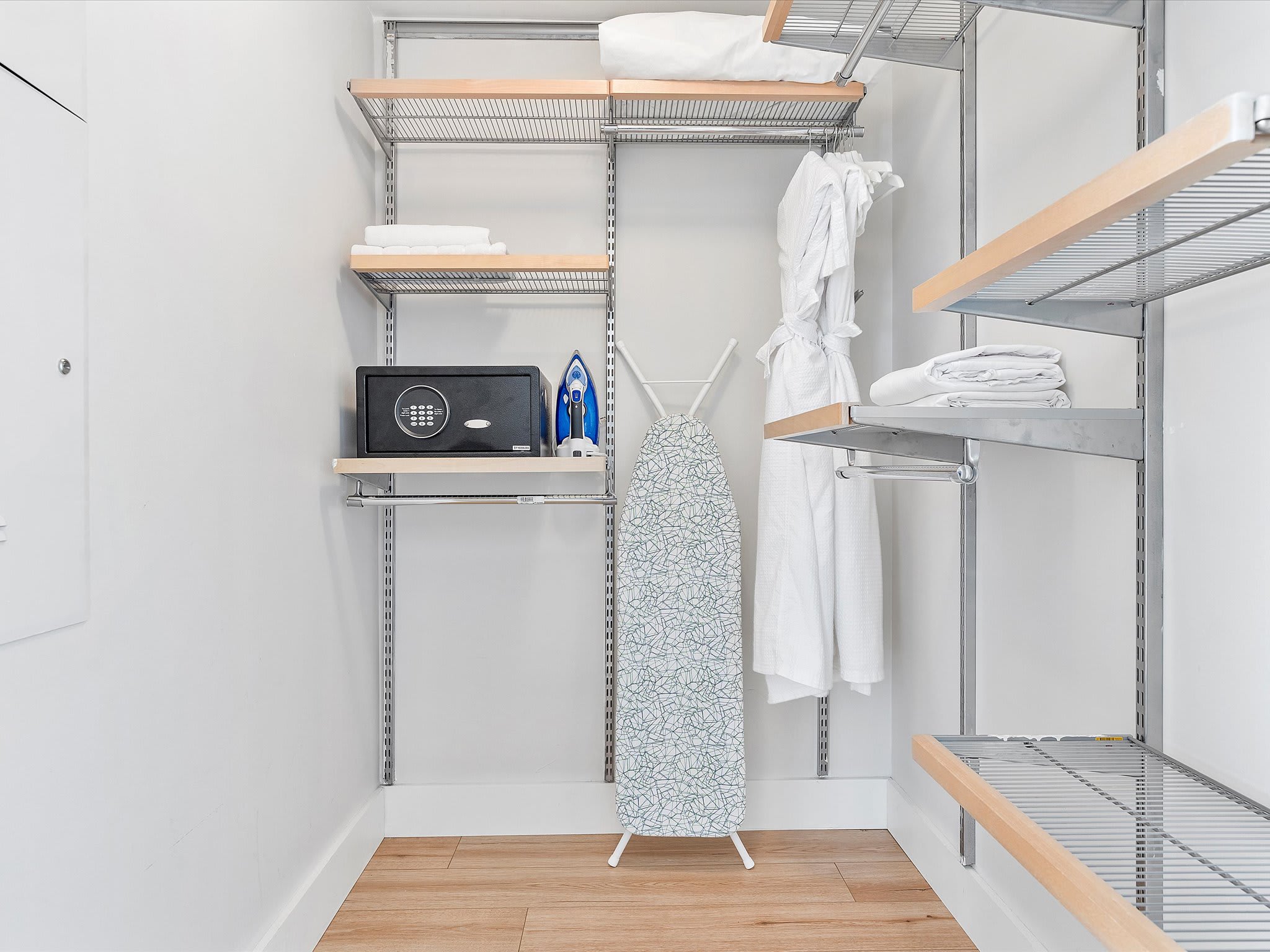 Organized walk-in closet featuring wire shelving units, a small safe, an ironing board, and neatly folded towels and bathrobes, providing ample storage space.