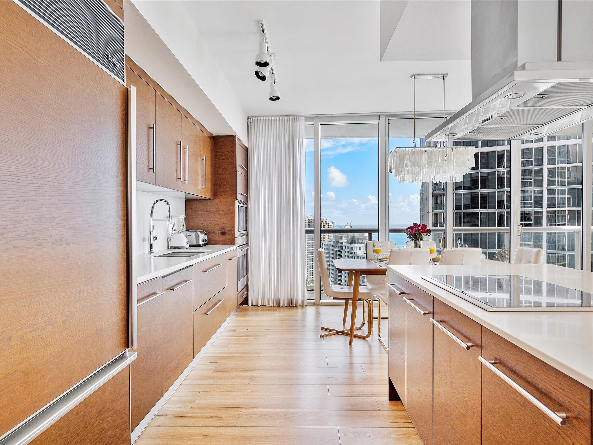 Sleek galley kitchen featuring light wood cabinets, white countertops, and integrated stainless steel appliances. The space opens to a dining area with floor-to-ceiling windows, offering bright natural light and city views.