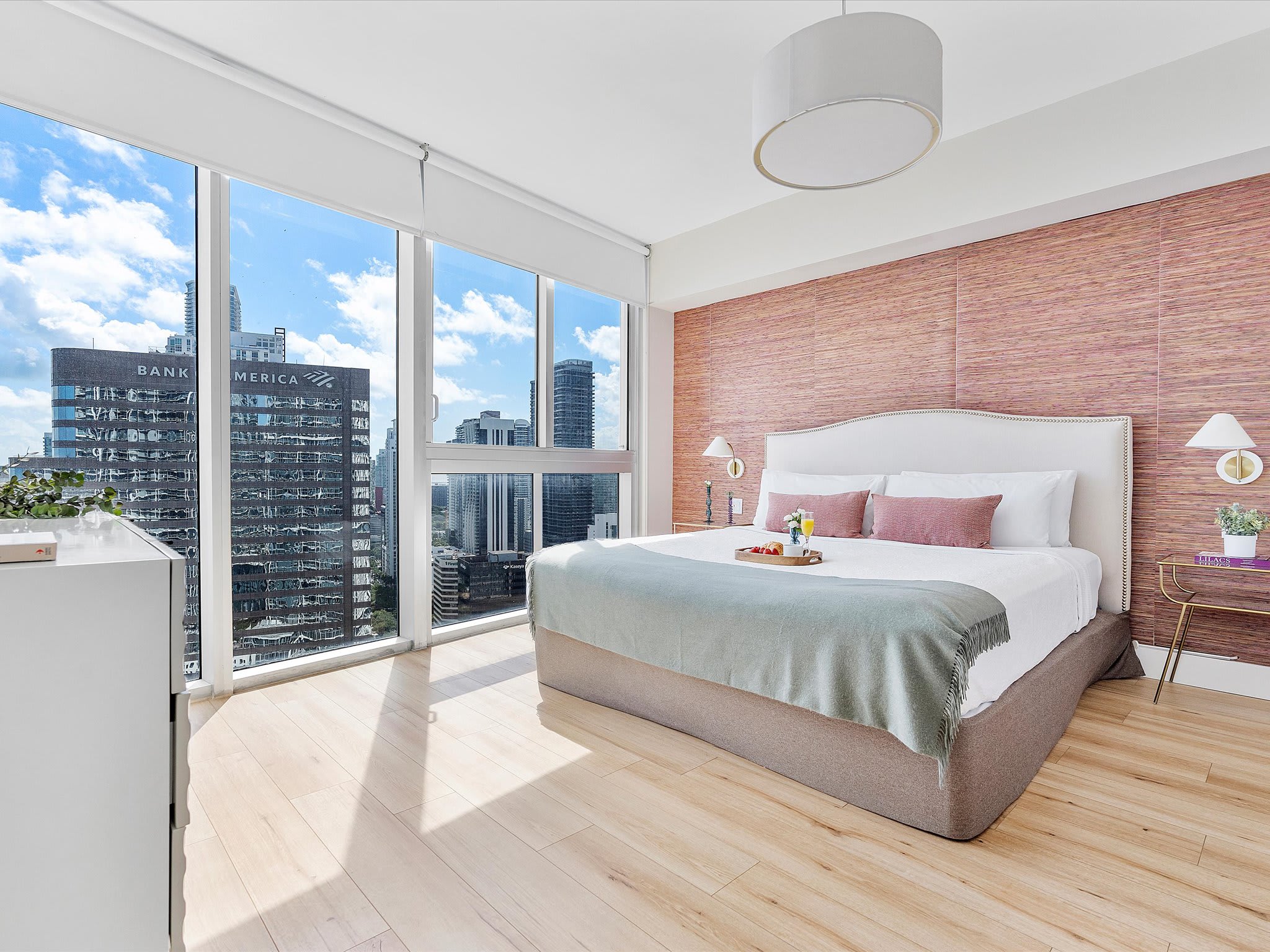 Bright primary bedroom featuring a king-sized bed with a white upholstered headboard and pink accents, light wood flooring, and floor-to-ceiling windows offering panoramic city views of downtown Miami.