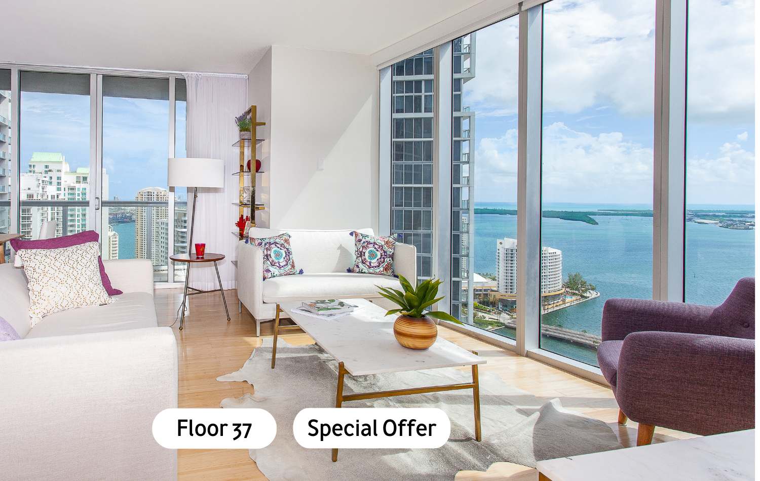 Bright living room with light hardwood floors, a white sofa, and a marble coffee table, featuring floor-to-ceiling windows offering panoramic bay and city views. A cowhide rug adds a touch of modern rustic style.
