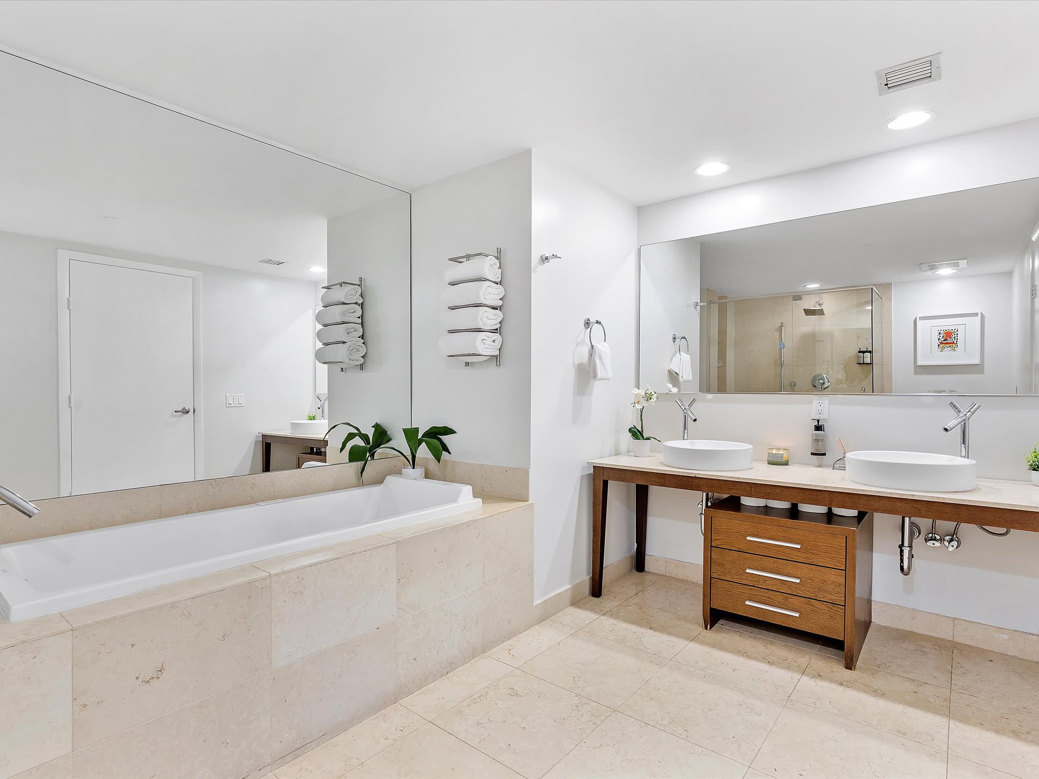 Spacious modern bathroom with light-colored tile flooring, a large built-in soaking tub, and a double vanity with vessel sinks and large mirrors, featuring clean white walls.