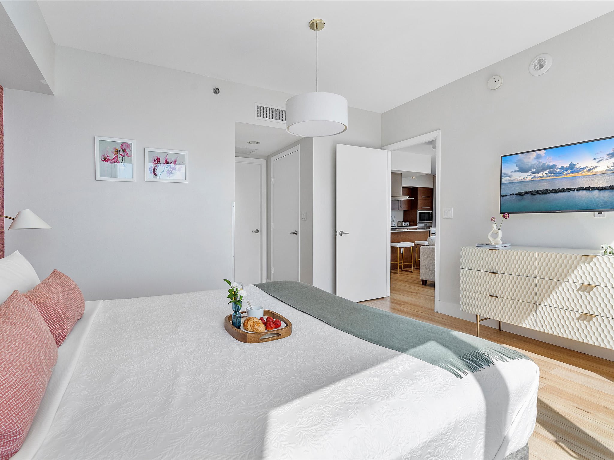 View of the primary bedroom with a king-sized bed, a white dresser with a flat-screen TV, and an open doorway leading to a kitchen area, featuring light wood floors and minimalist decor.