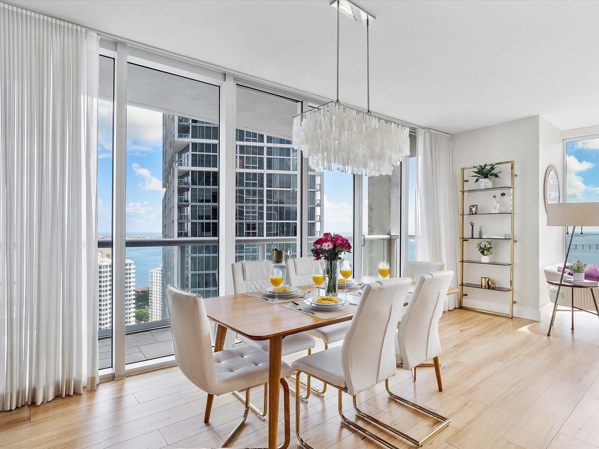 Elegant dining area with a light wood table and six white upholstered chairs, illuminated by a modern rectangular chandelier. Floor-to-ceiling windows provide expansive city and bay views, with access to a balcony.