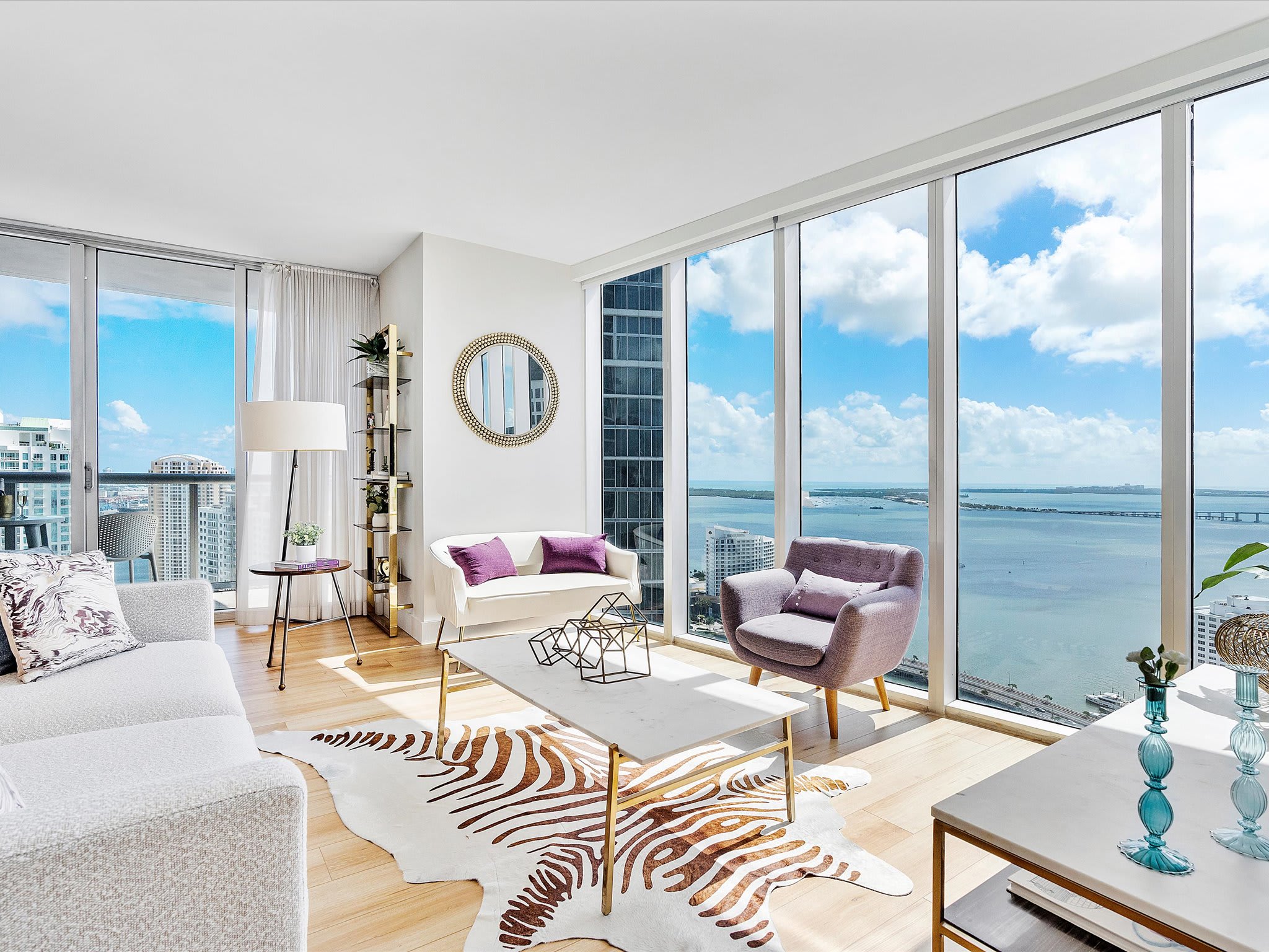 Spacious living room with light hardwood floors, a large white sectional sofa, and a marble coffee table over a zebra-print rug. Floor-to-ceiling windows offer expansive bay and city views, complemented by a round gold-framed mirror.