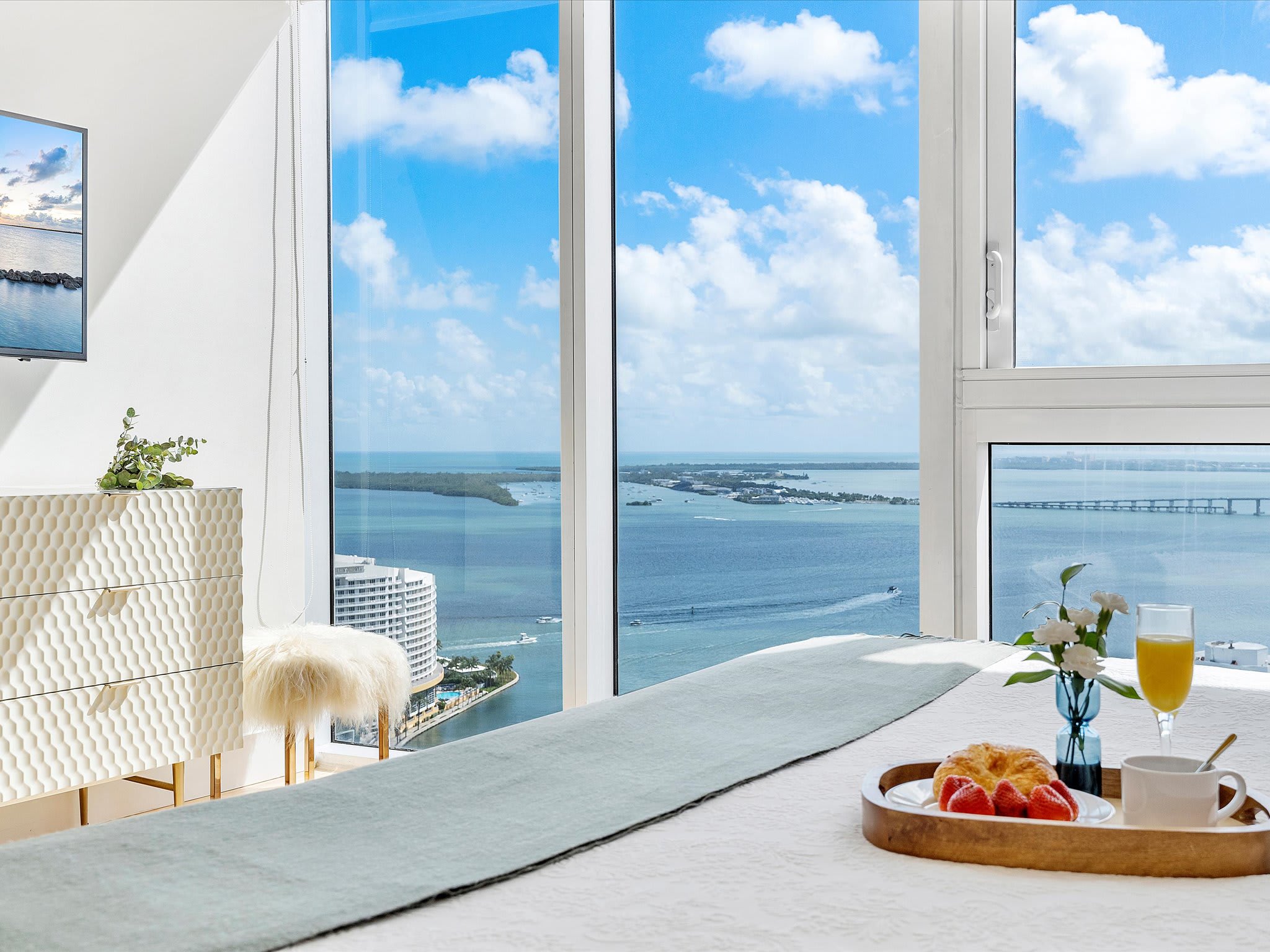Bright bedroom with a white bed and a textured dresser, featuring floor-to-ceiling windows that provide stunning bay and city views. A breakfast tray with fruit and juice is placed on the bed, suggesting a comfortable stay.