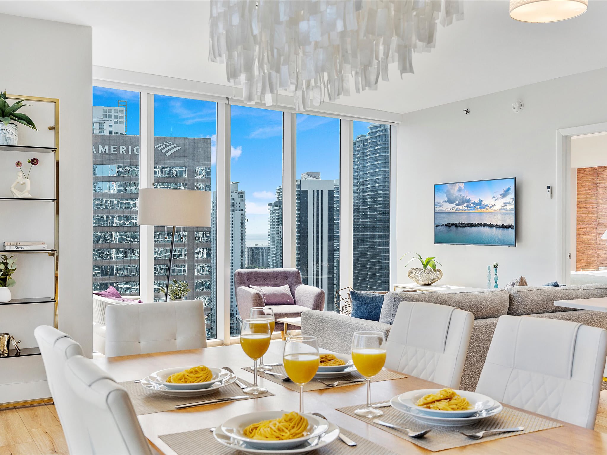 Elegant dining area with a light wood table set for four, white upholstered chairs, and a striking modern chandelier. Floor-to-ceiling windows provide a city view, and a wall-mounted TV is visible in the adjacent living space.