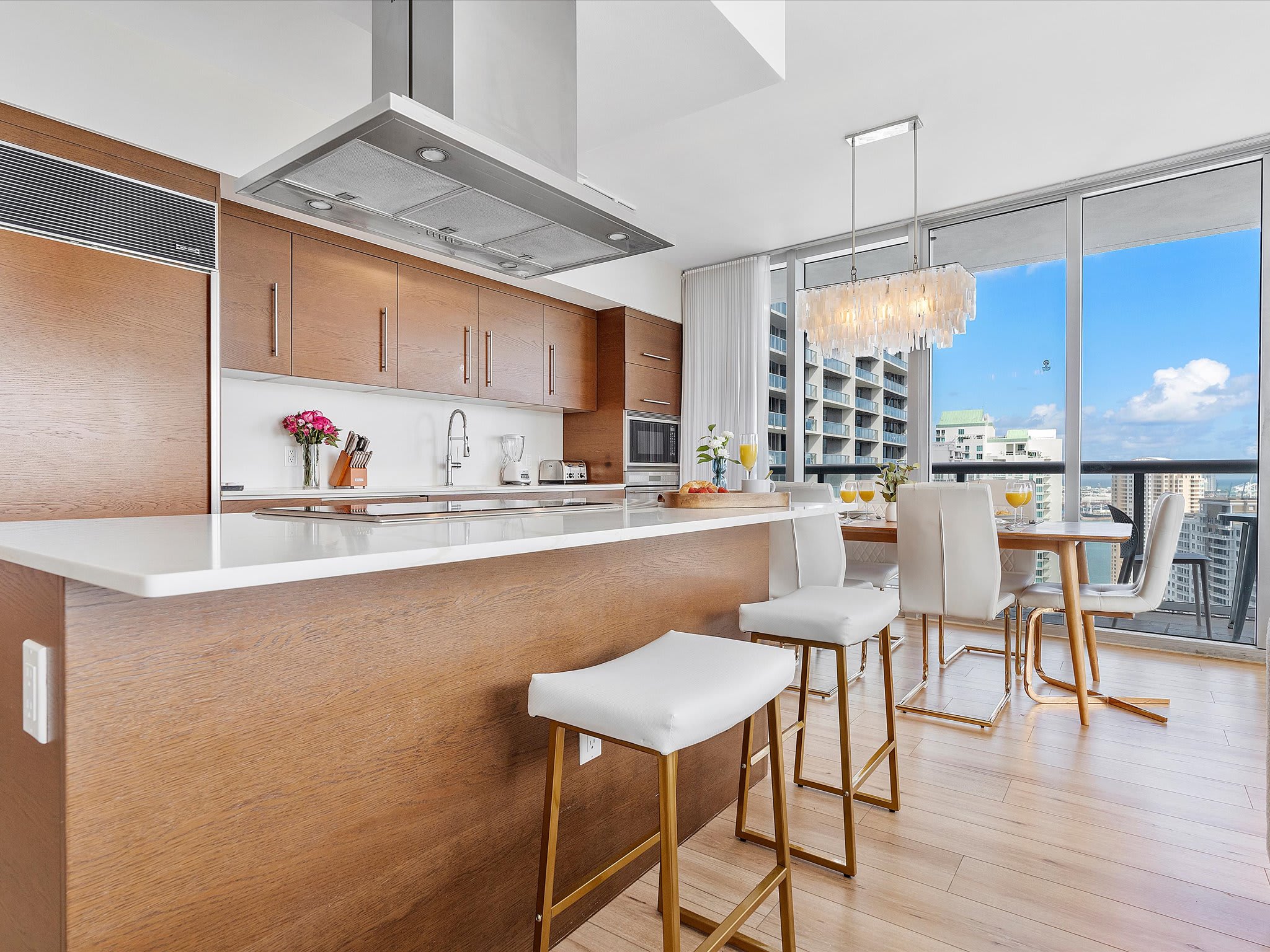 Modern kitchen with light wood cabinetry, white countertops, and stainless steel appliances, including a large island with bar seating. The open-concept space flows into a dining area with floor-to-ceiling windows and city views.