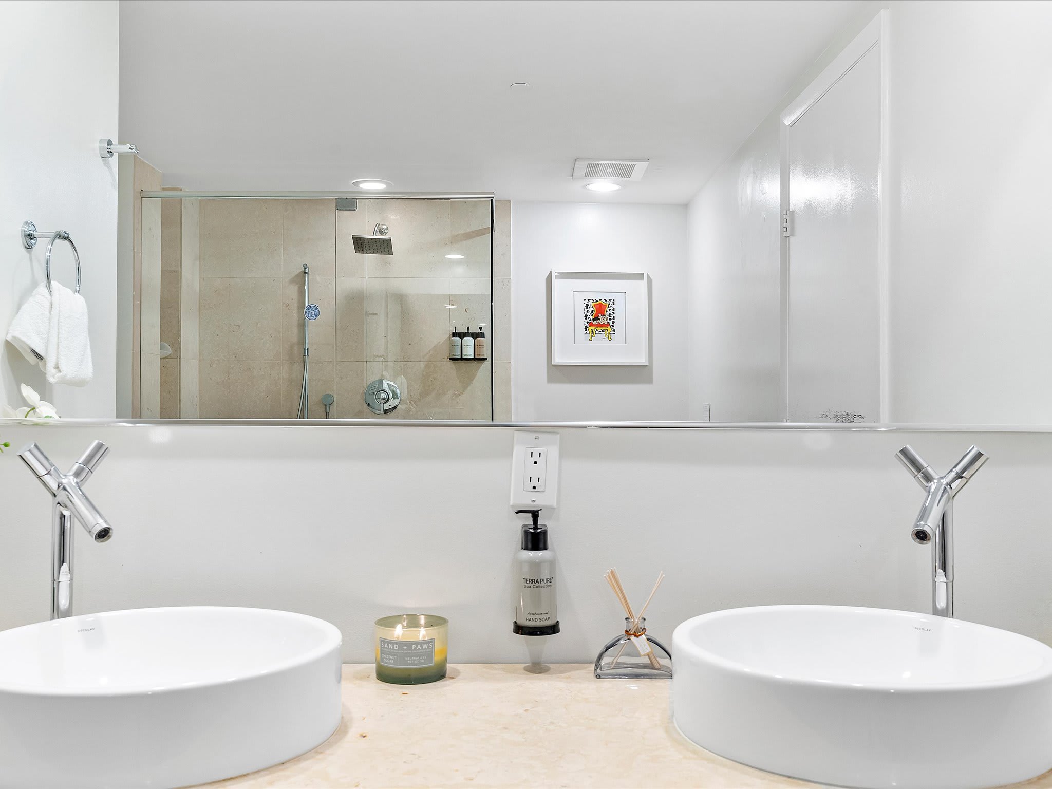 Contemporary double vanity bathroom with two white vessel sinks, chrome faucets, and a large mirror reflecting the walk-in shower. A soap dispenser and a decorative candle are placed between the sinks.