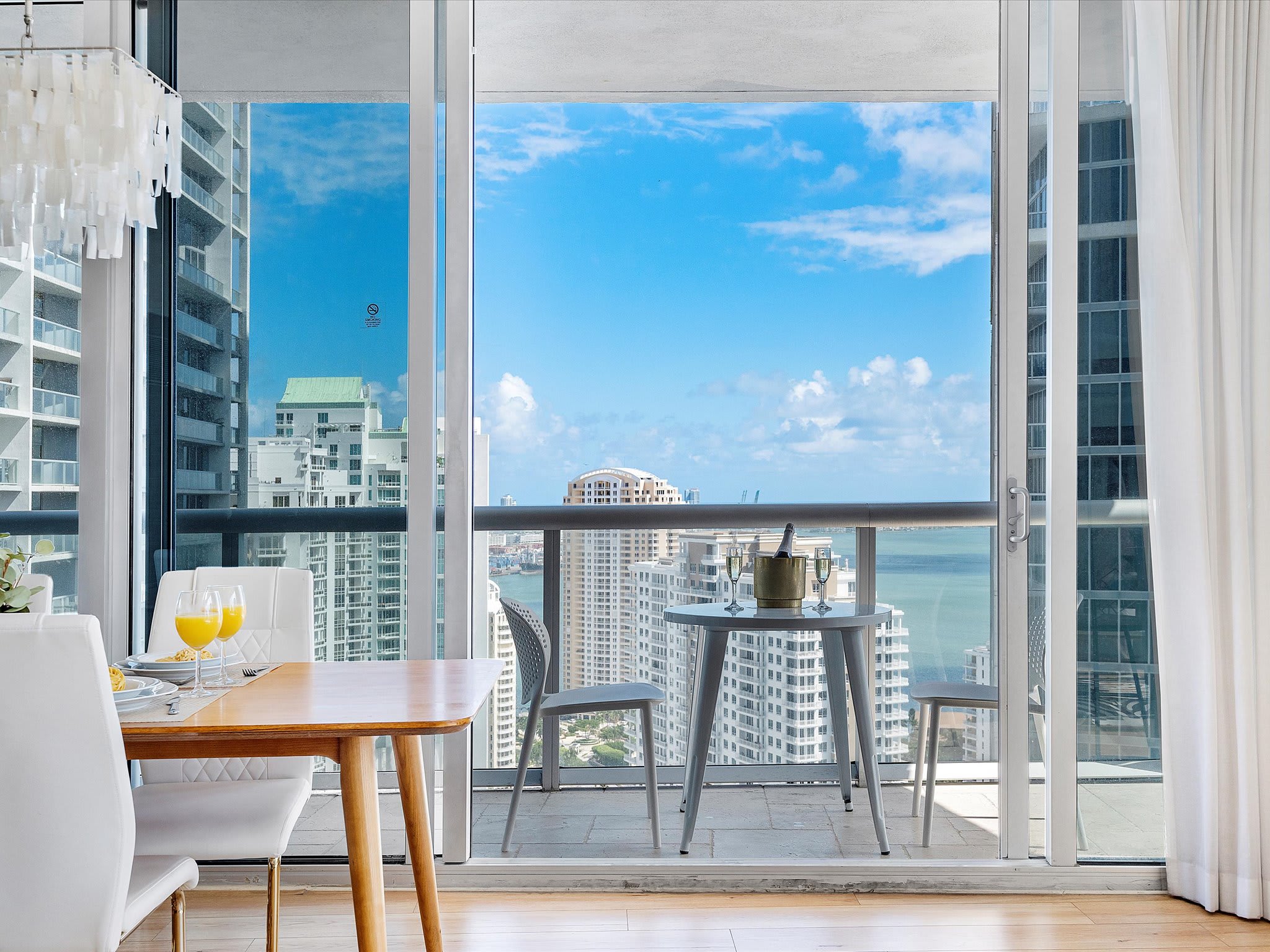 Modern dining area with a light wood table and white chairs, featuring a large sliding glass door leading to a balcony with a small bistro set and expansive bay and city views.