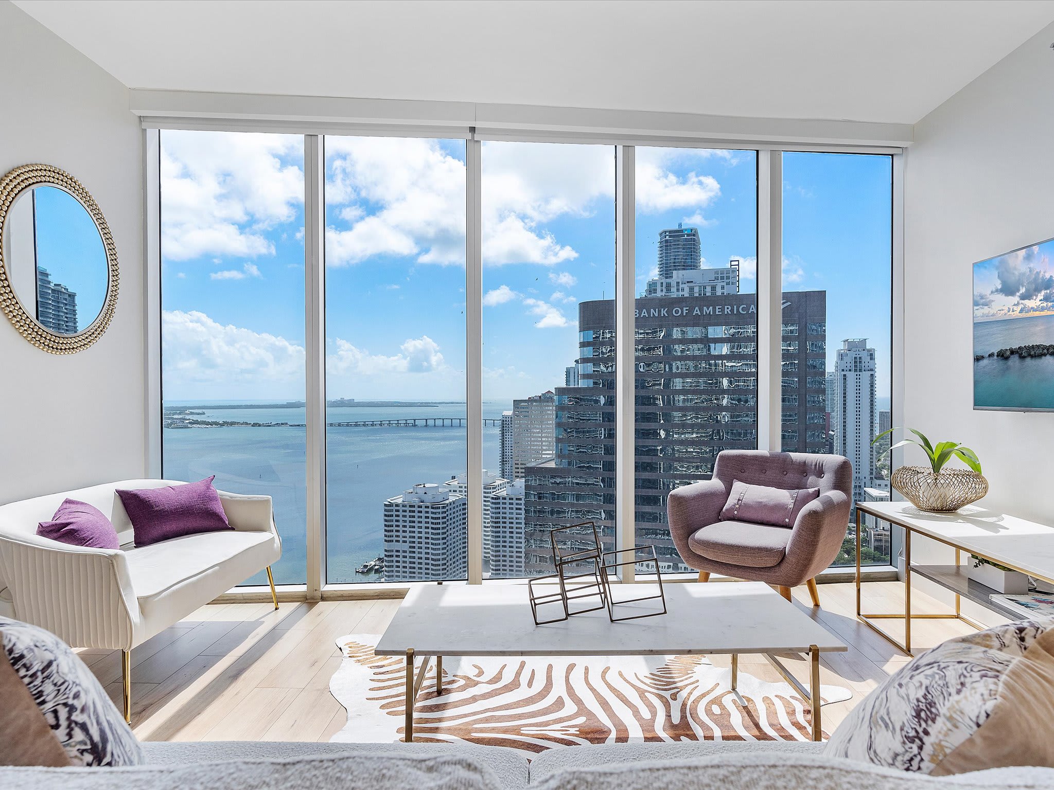 Modern living room with a white sofa and a purple accent chair, set on light hardwood floors with a zebra-print rug. Floor-to-ceiling windows provide breathtaking city and bay views, with a large building visible outside.