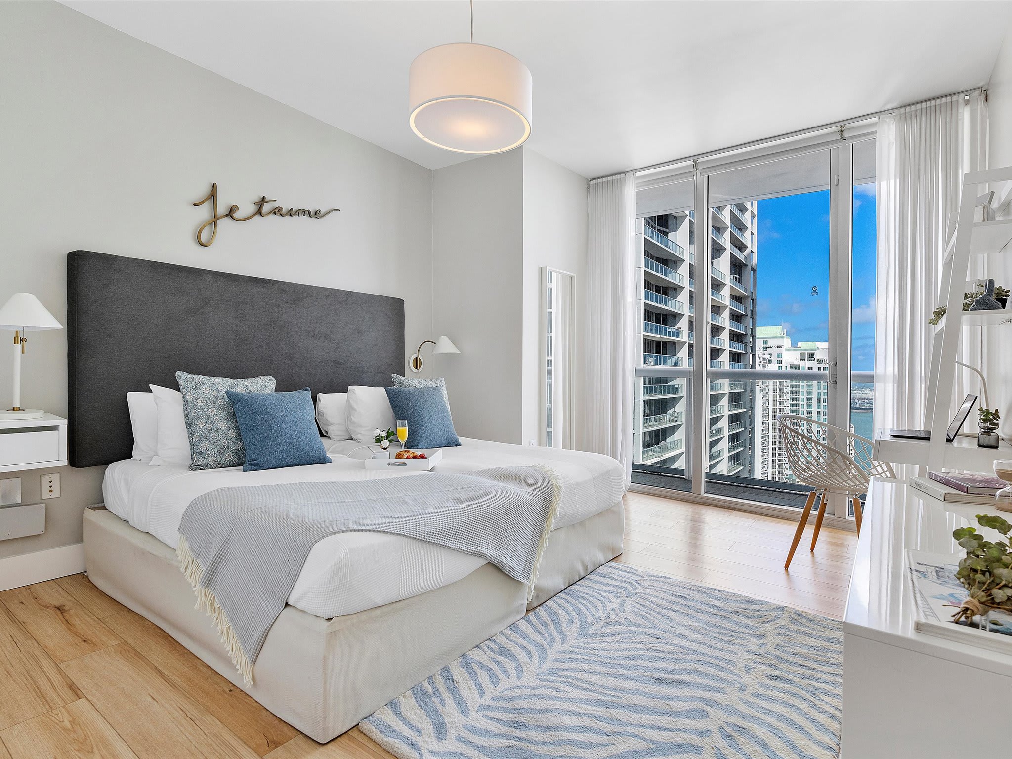 Second bedroom featuring a king-sized bed with a dark grey upholstered headboard, a blue and white patterned rug, and large windows offering city and partial bay views.