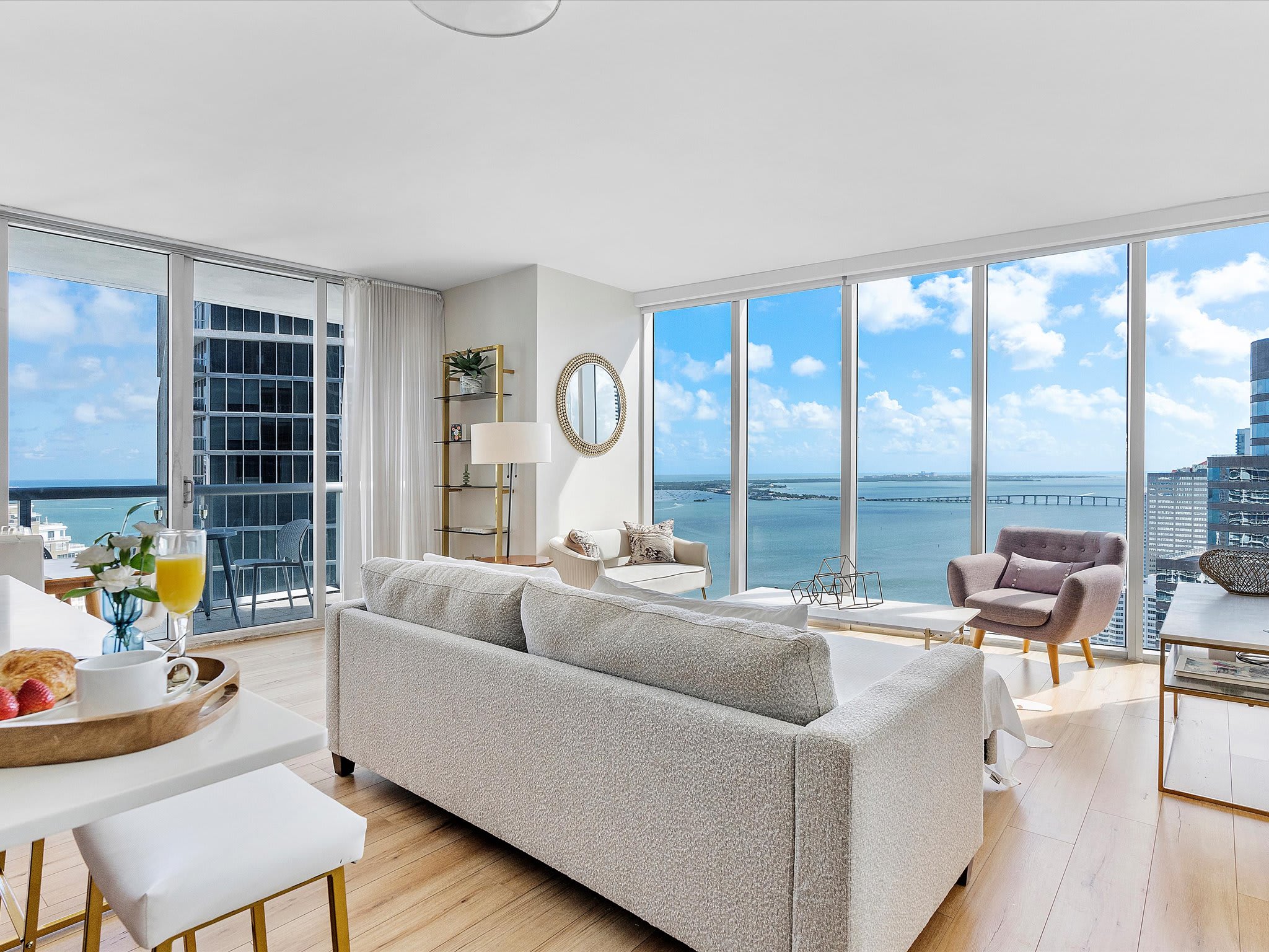 Bright and airy living room with a large white sofa and a marble coffee table, featuring light hardwood floors. Floor-to-ceiling windows offer stunning bay and city views, with a glimpse of the adjacent dining area.