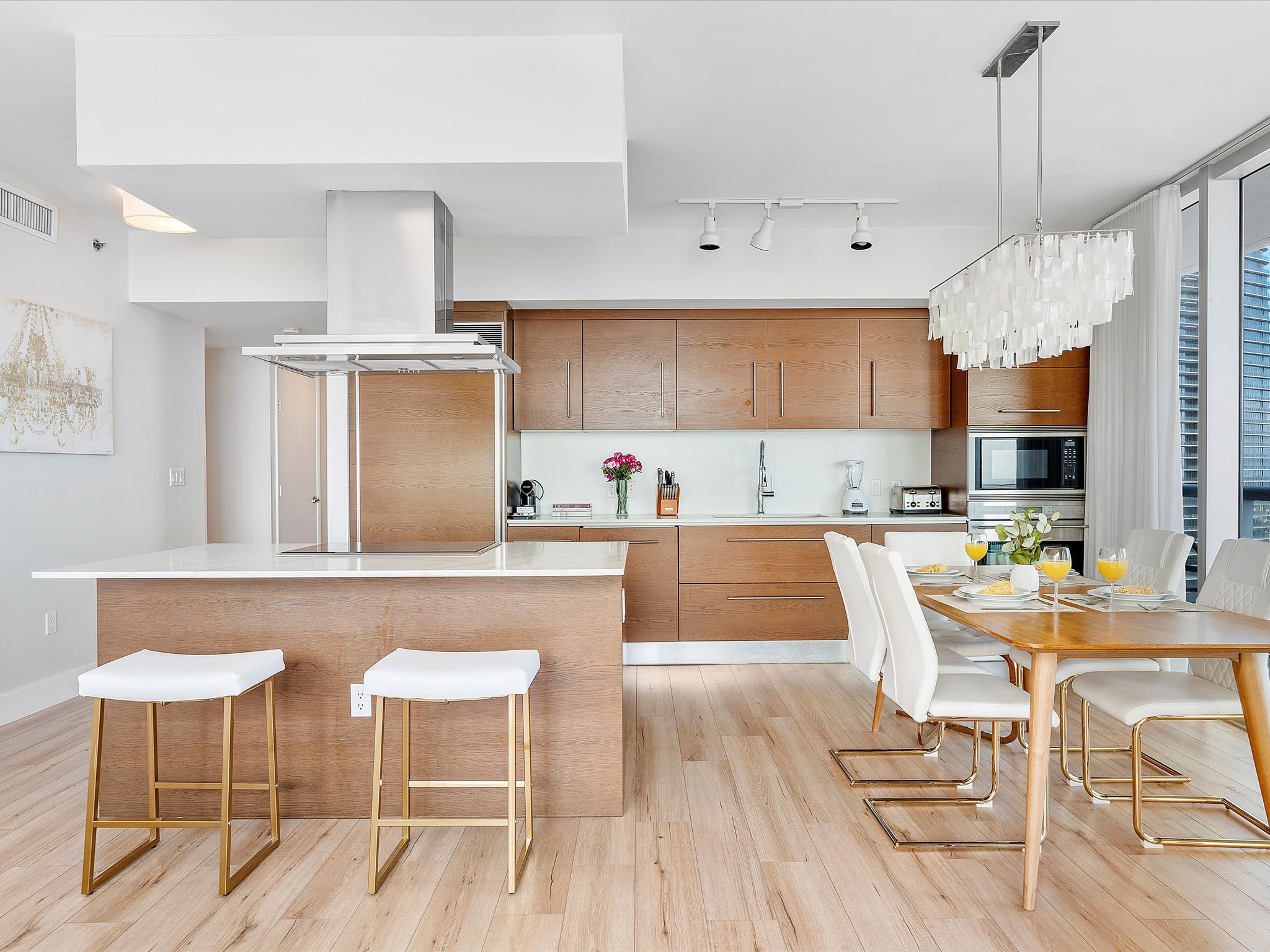 Open-concept kitchen and dining area with light wood cabinetry, white countertops, and a large island with two white bar stools. The dining table is set for four, and the space is brightly lit by natural light from floor-to-ceiling windows.