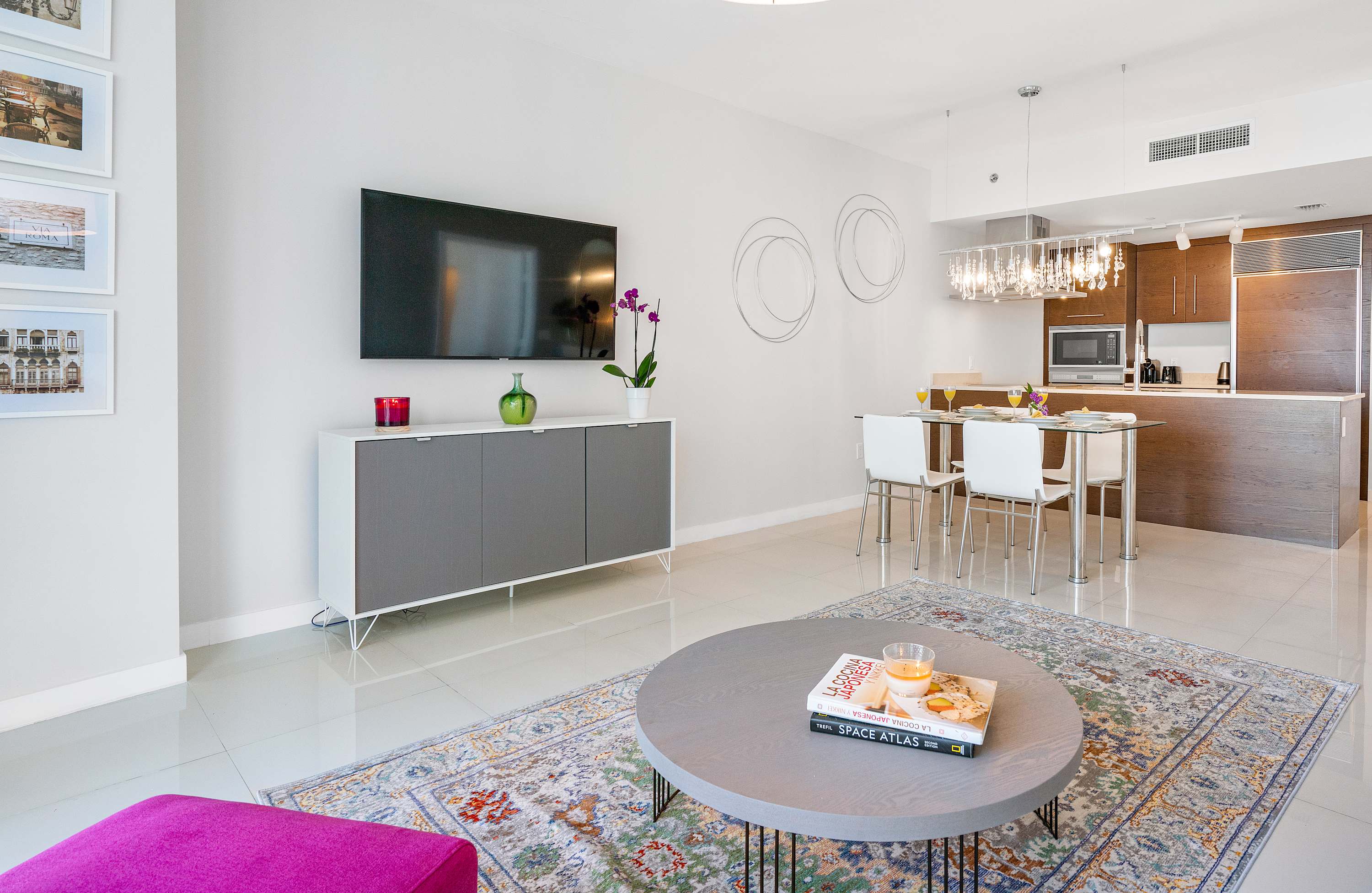 A bright, contemporary living room features a wall-mounted flat-screen TV above a gray and white media console. A round wooden coffee table sits on a colorful patterned rug, with a vibrant fuchsia ottoman in the foreground.