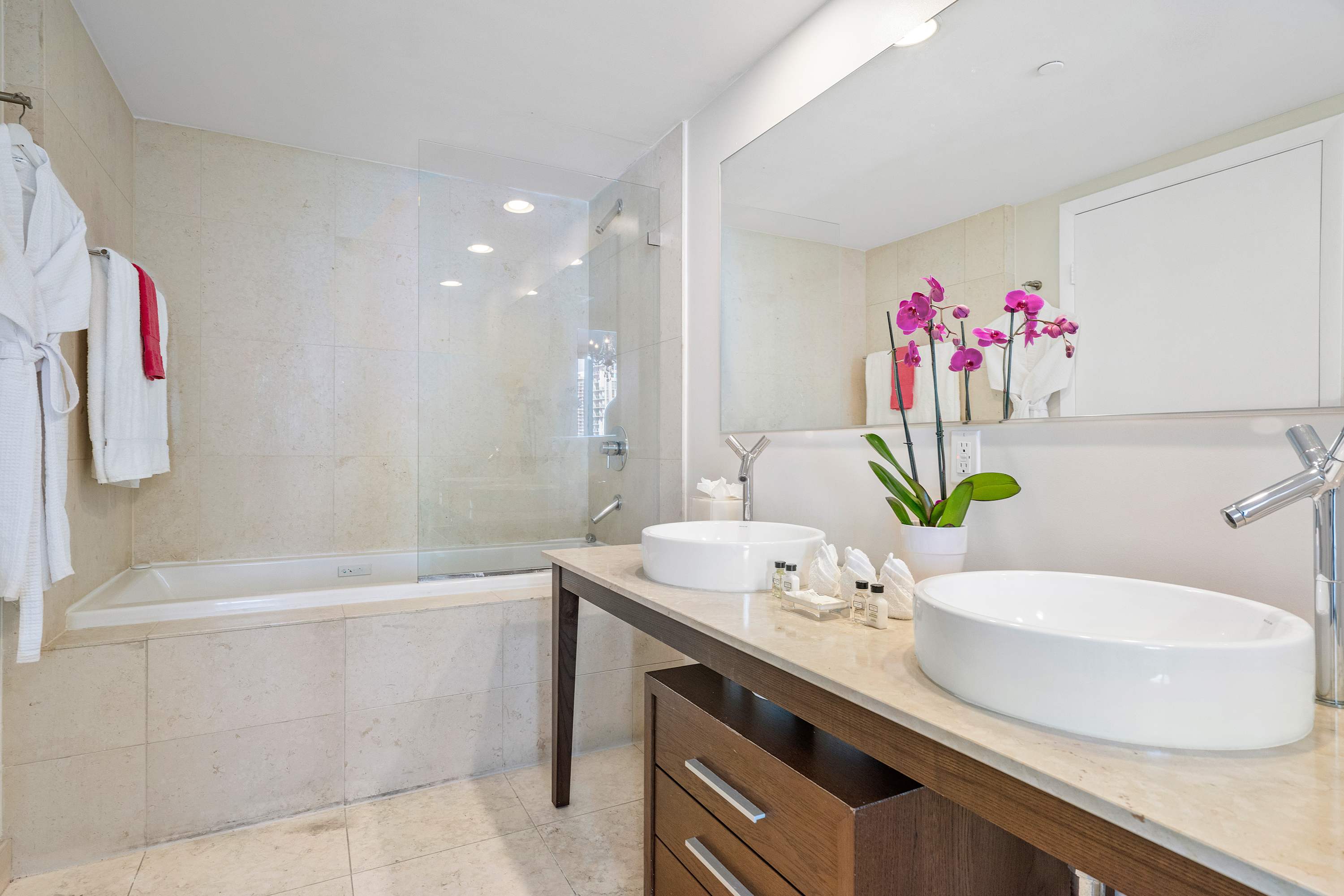 A bright bathroom showcases a double vanity with white vessel sinks and modern fixtures, alongside a large mirror. A tiled bathtub and shower area with a glass screen are visible, with white bathrobes hanging nearby.