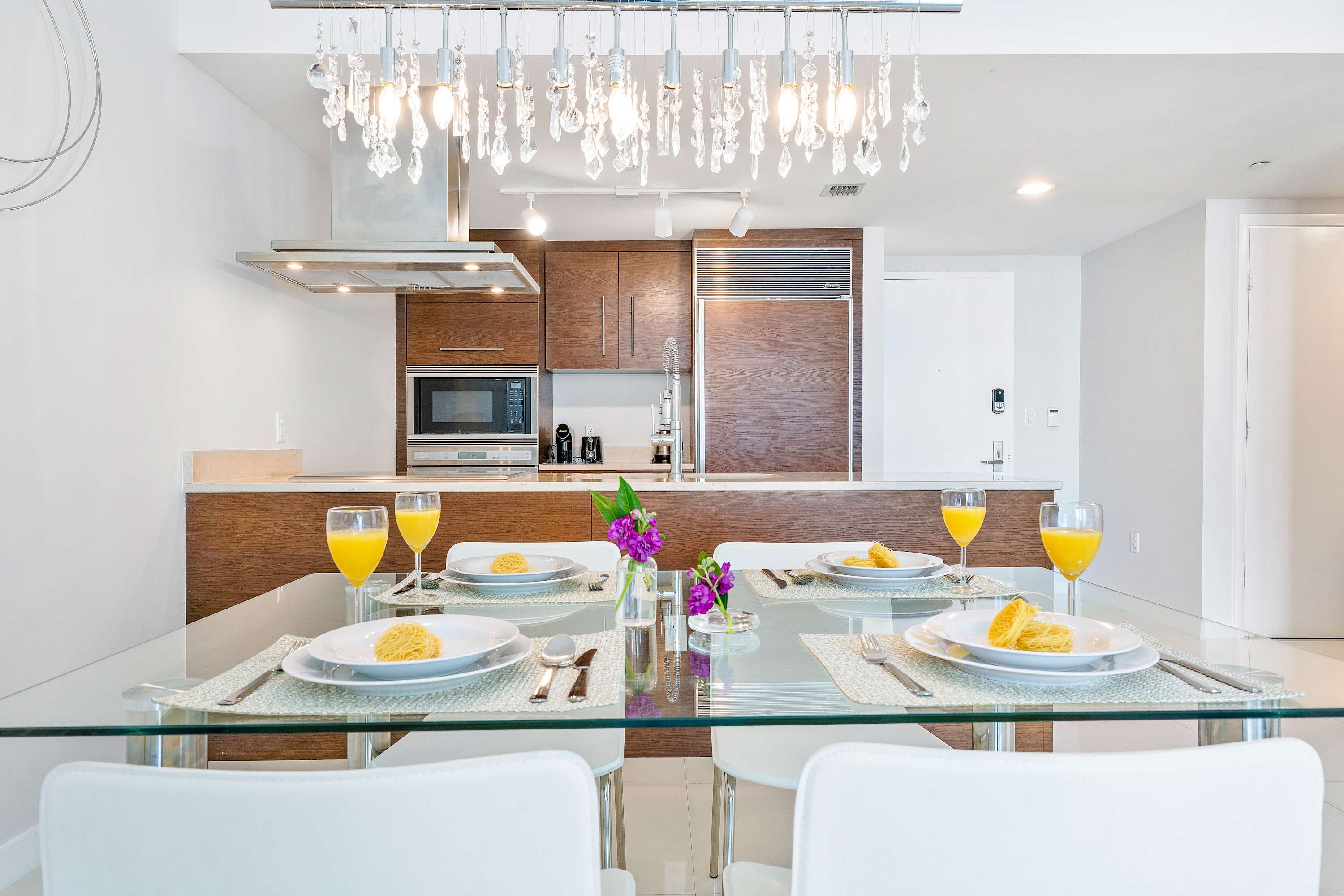 A modern dining area features a clear glass table set for four with white chairs, illuminated by a crystal chandelier. In the background, a sleek kitchen with dark wood cabinets, stainless steel appliances, and a light countertop is visible.
