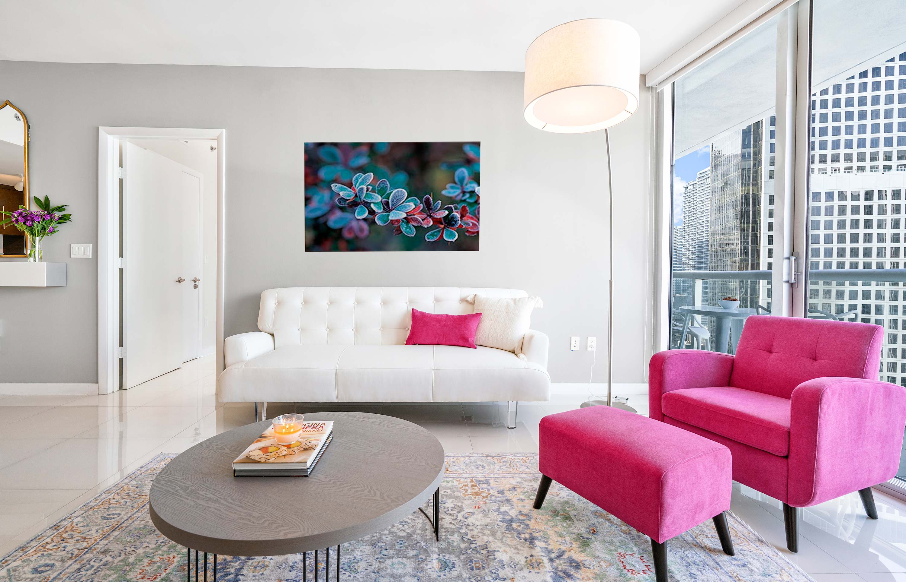 A contemporary living room features a white two-seater sofa, a vibrant pink armchair with an ottoman, and a round wooden coffee table, all resting on a patterned area rug, with large windows offering city views.