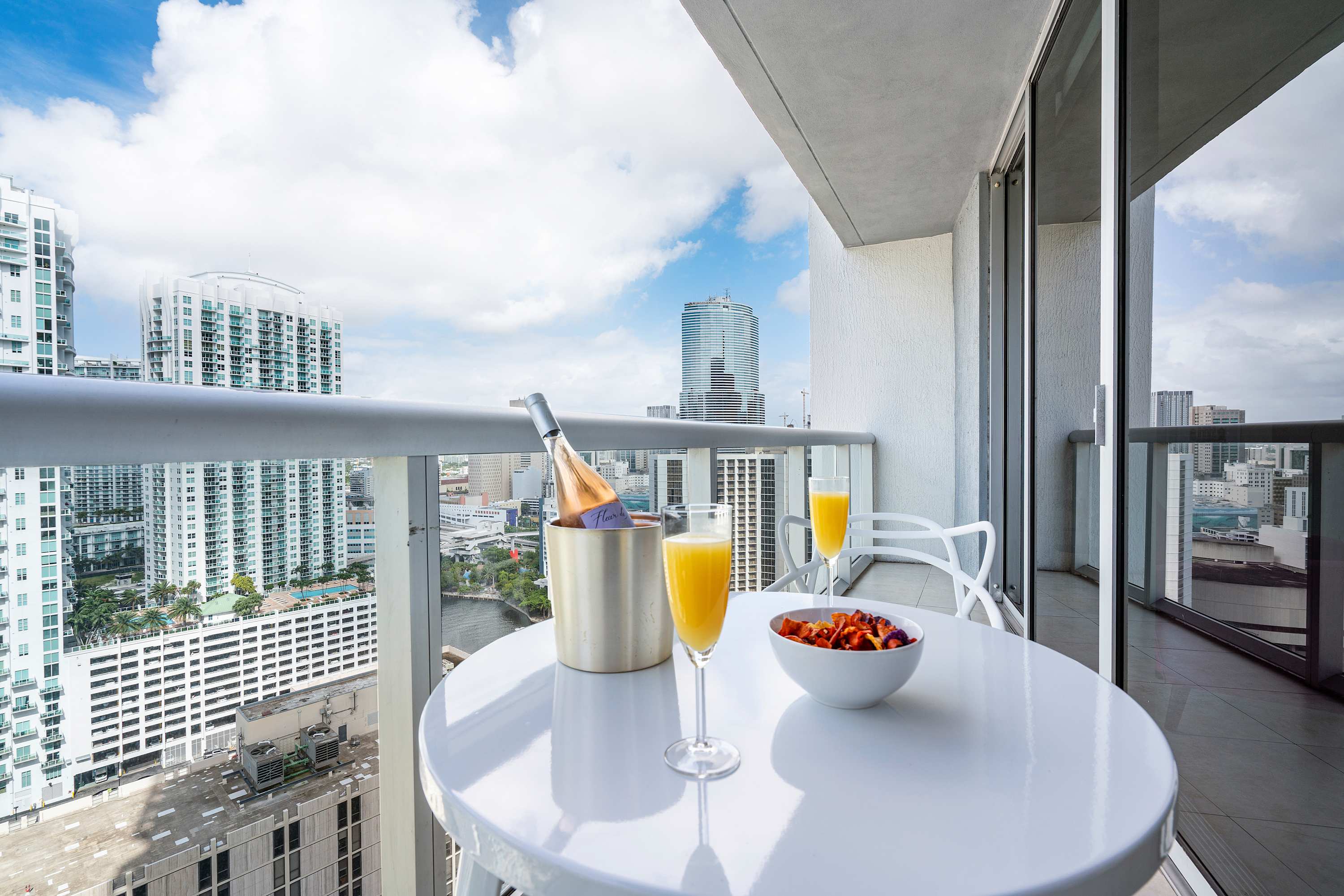 A high-rise balcony offers expansive city and waterfront views, featuring a white outdoor table with two chairs, a bottle of sparkling wine in a cooler, two mimosas, and a bowl of snacks.