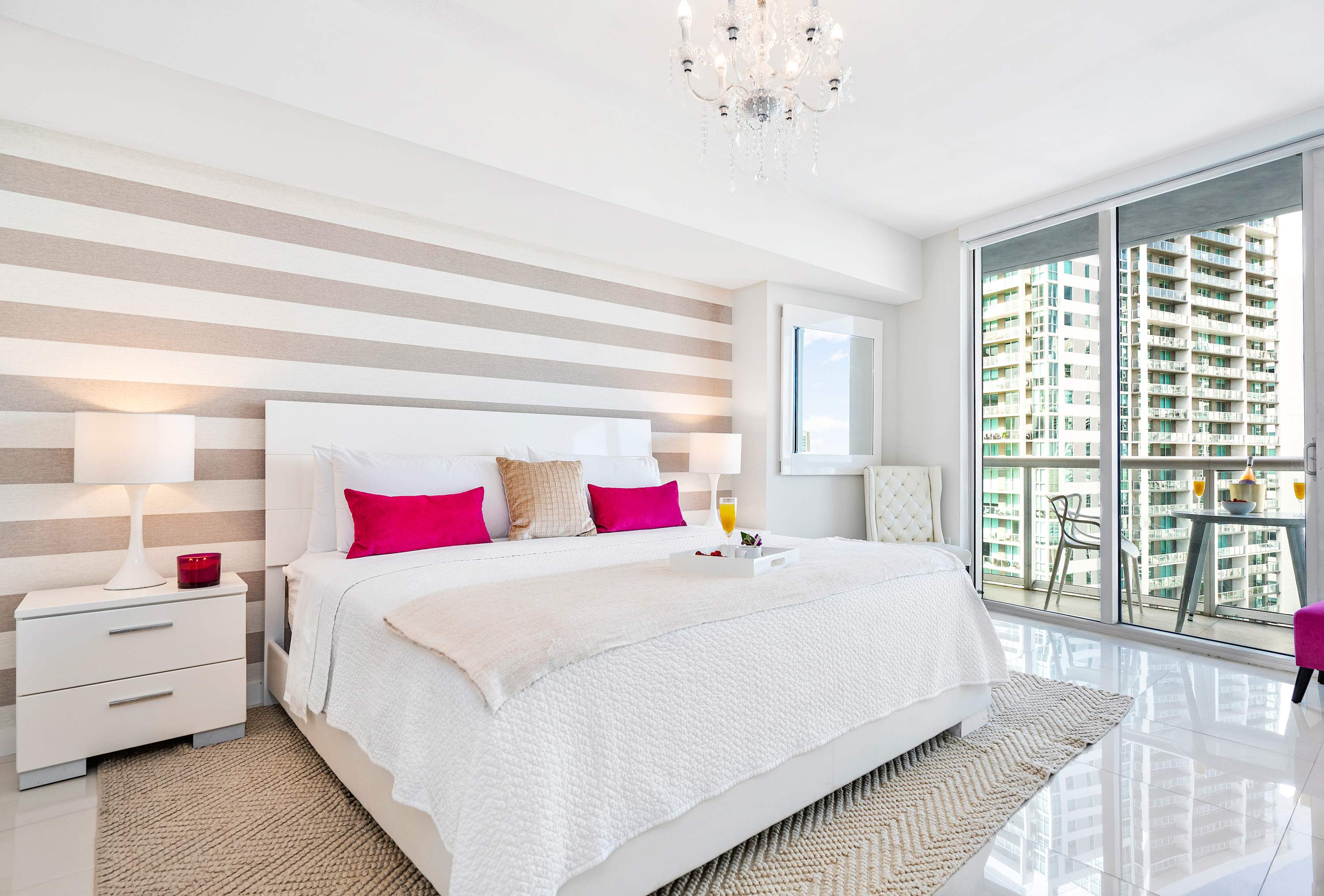 A spacious bedroom features a white king-sized bed with fuchsia and gold accent pillows, set against a striped accent wall. A crystal chandelier hangs from the ceiling, and large glass doors open to a balcony with city views.