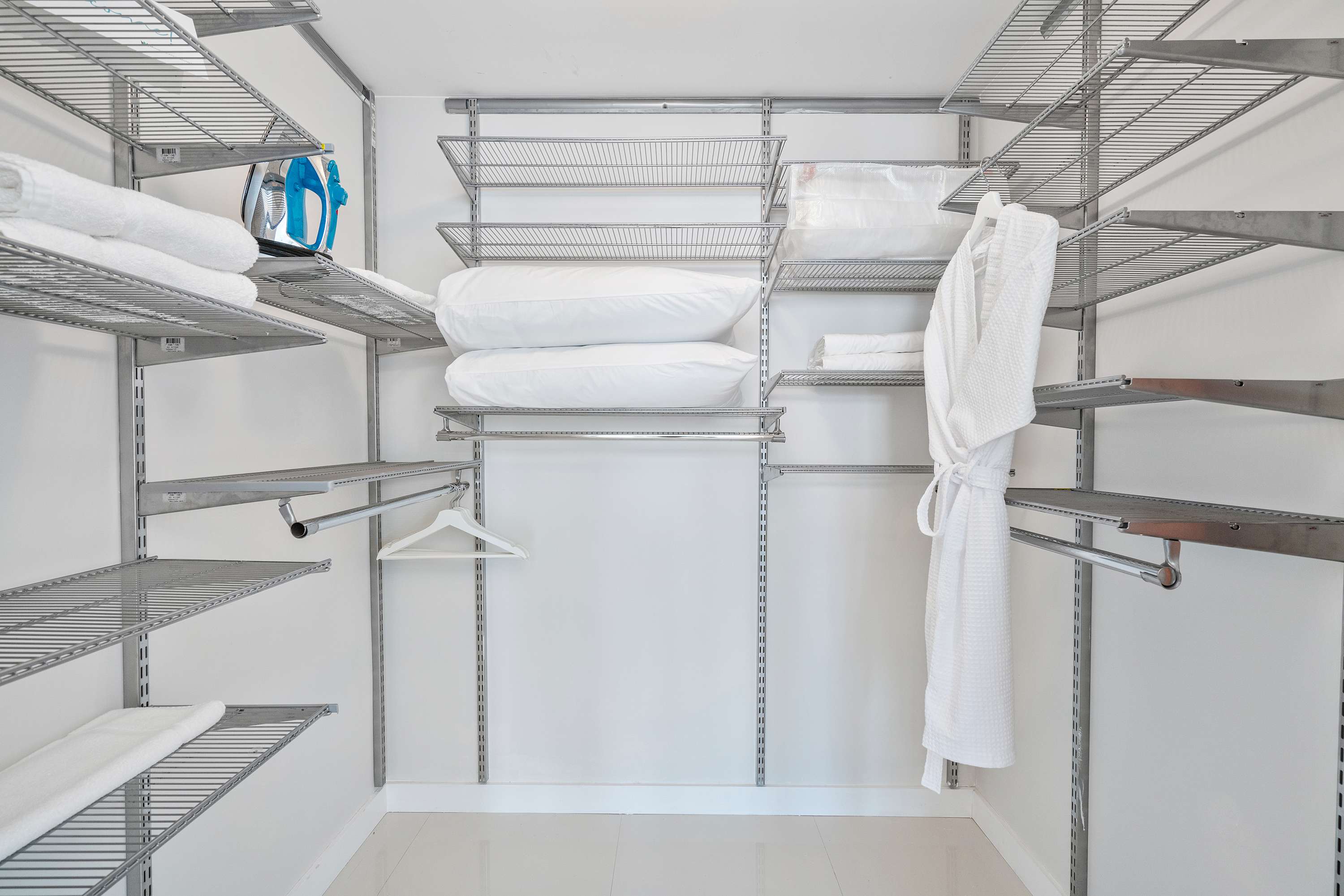 A well-organized walk-in closet features extensive wire shelving systems on all walls, providing ample storage space. White towels, pillows, an iron, and a bathrobe are neatly stored on the shelves.