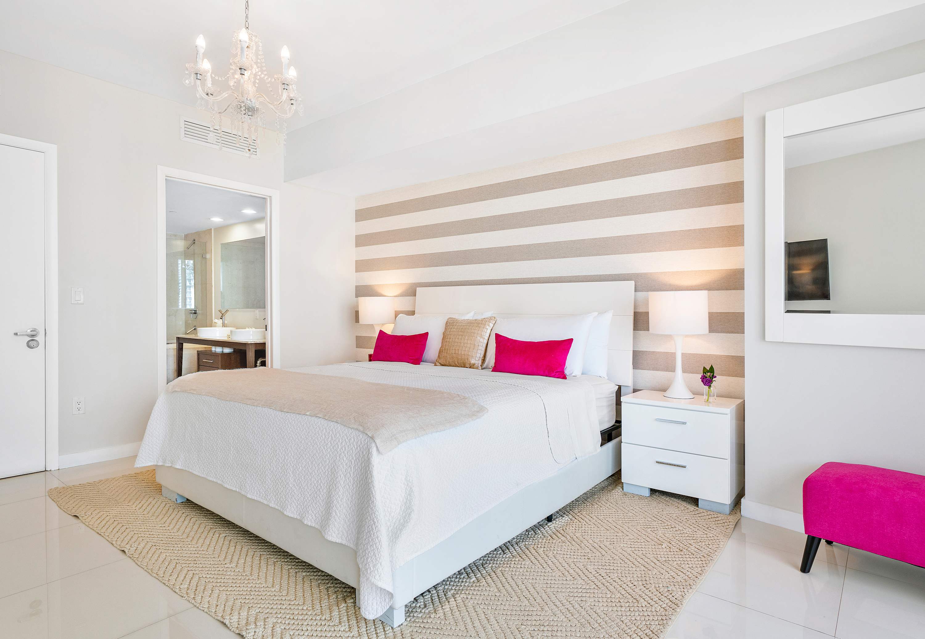 A spacious bedroom features a king-sized bed with a white headboard and a striped accent wall, flanked by white nightstands and lamps, with a crystal chandelier overhead and an open doorway leading to an en-suite bathroom.