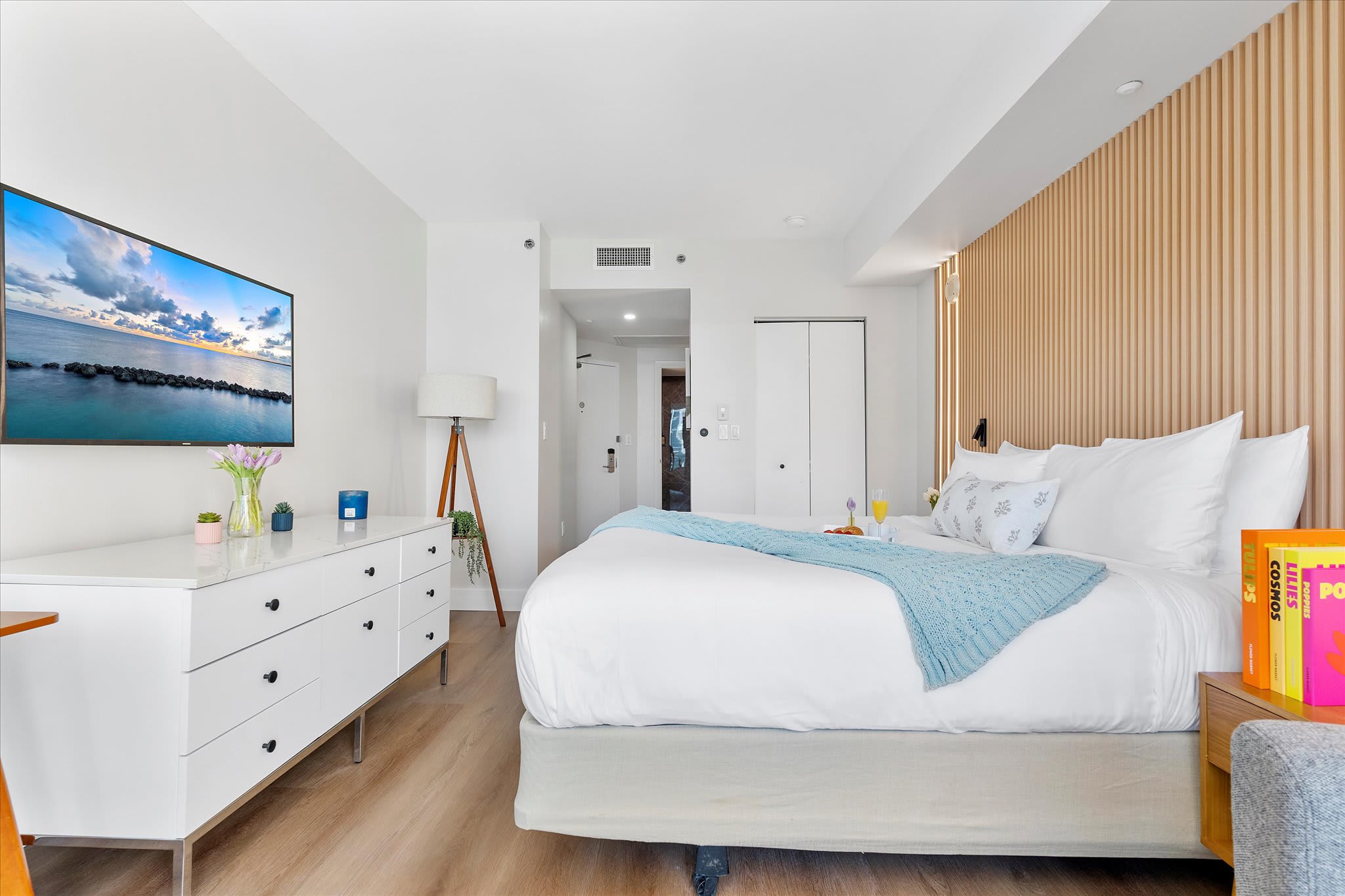 A wide shot of the modern bedroom shows a king bed with a light blue throw, a white dresser with a mounted flat-screen TV, and a wooden slatted accent wall behind the bed. A floor lamp and closet doors are visible.