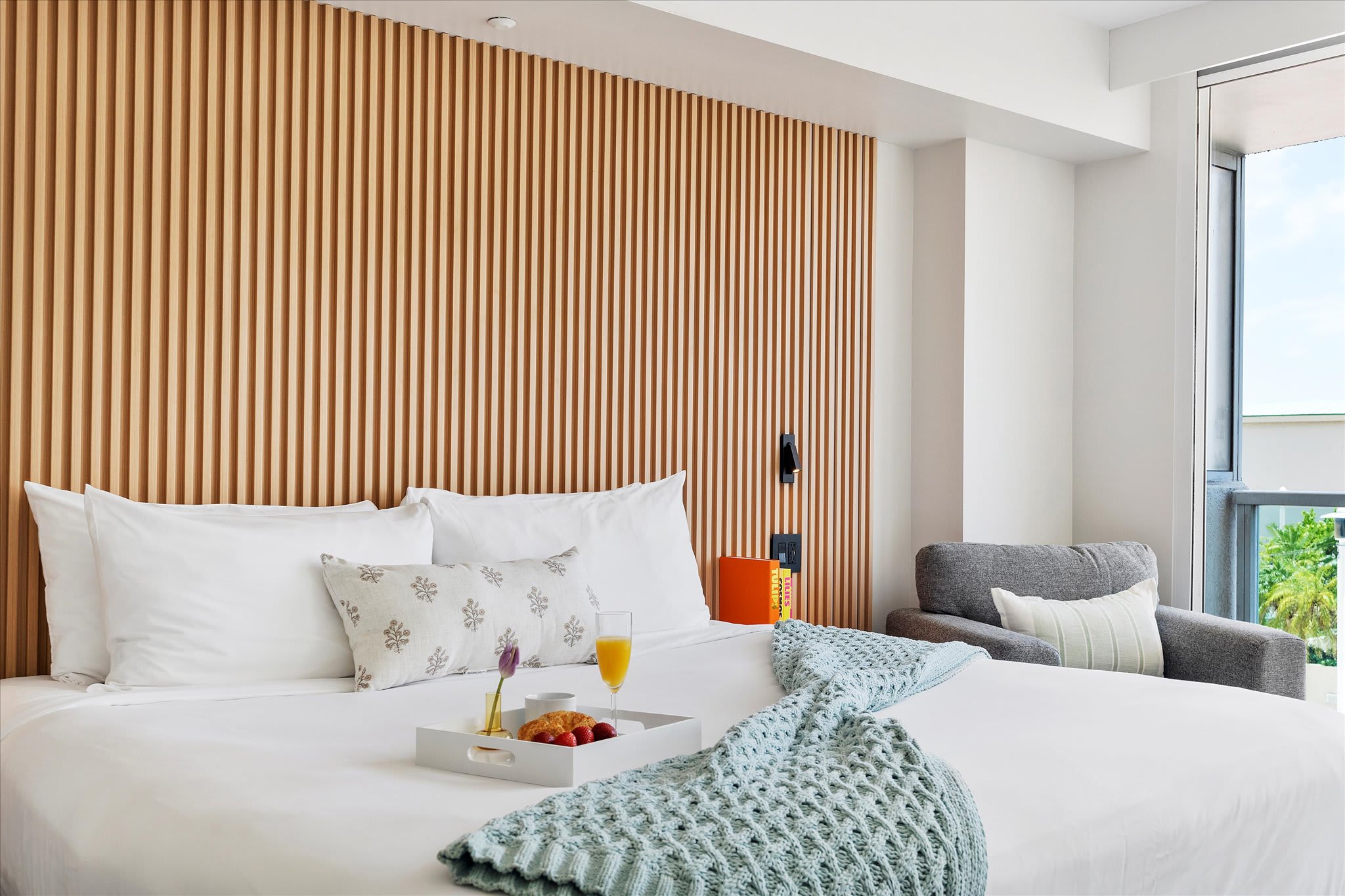 A modern bedroom features a king-sized bed with white linens and a light blue knitted throw, set against a slatted wooden accent wall, with a grey armchair and large window to the right. A breakfast tray with fruit and juice rests on the bed.