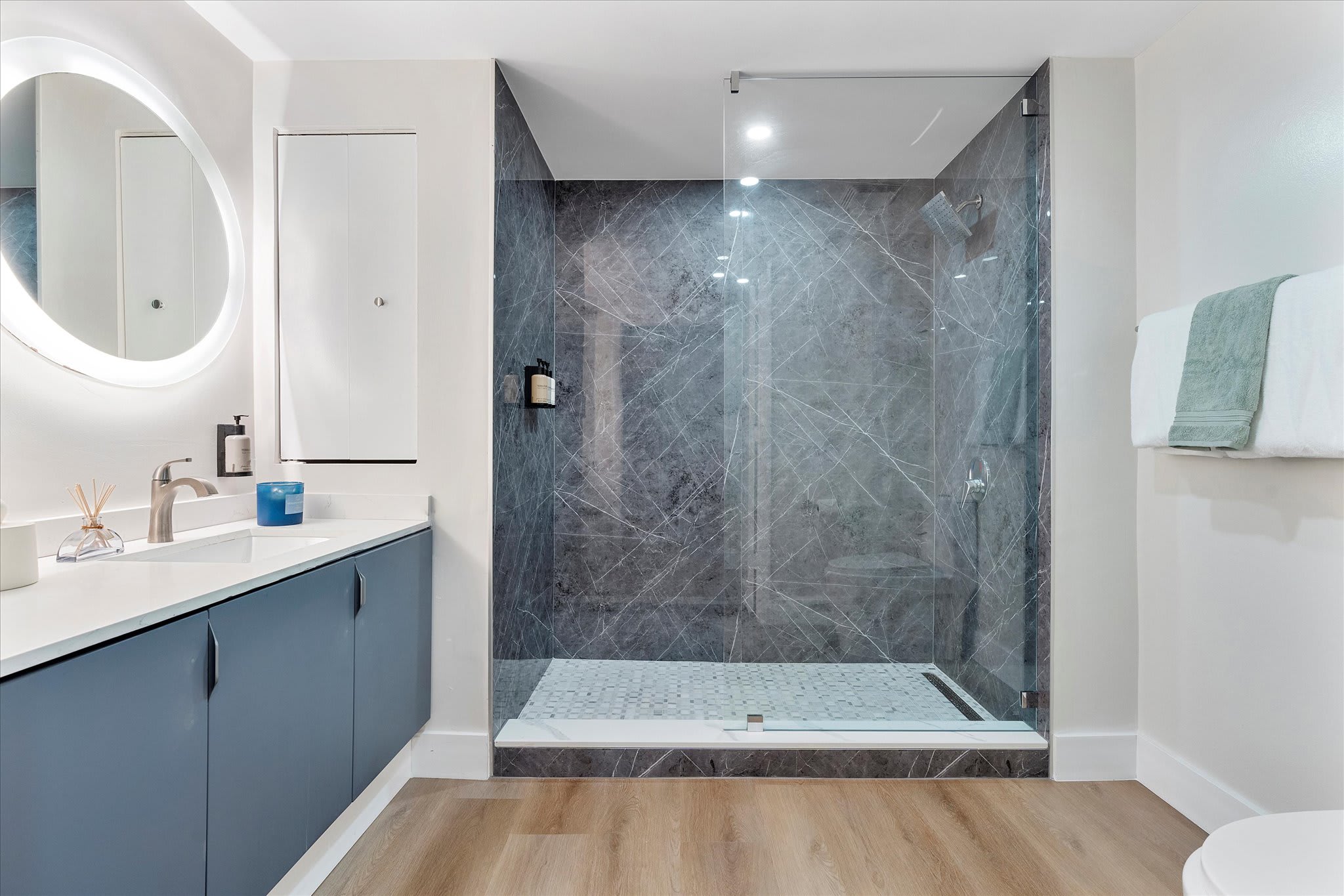 A contemporary bathroom features a large walk-in shower with dark grey marble-patterned tiles and a glass enclosure, alongside a vanity with a blue cabinet and a round illuminated mirror.