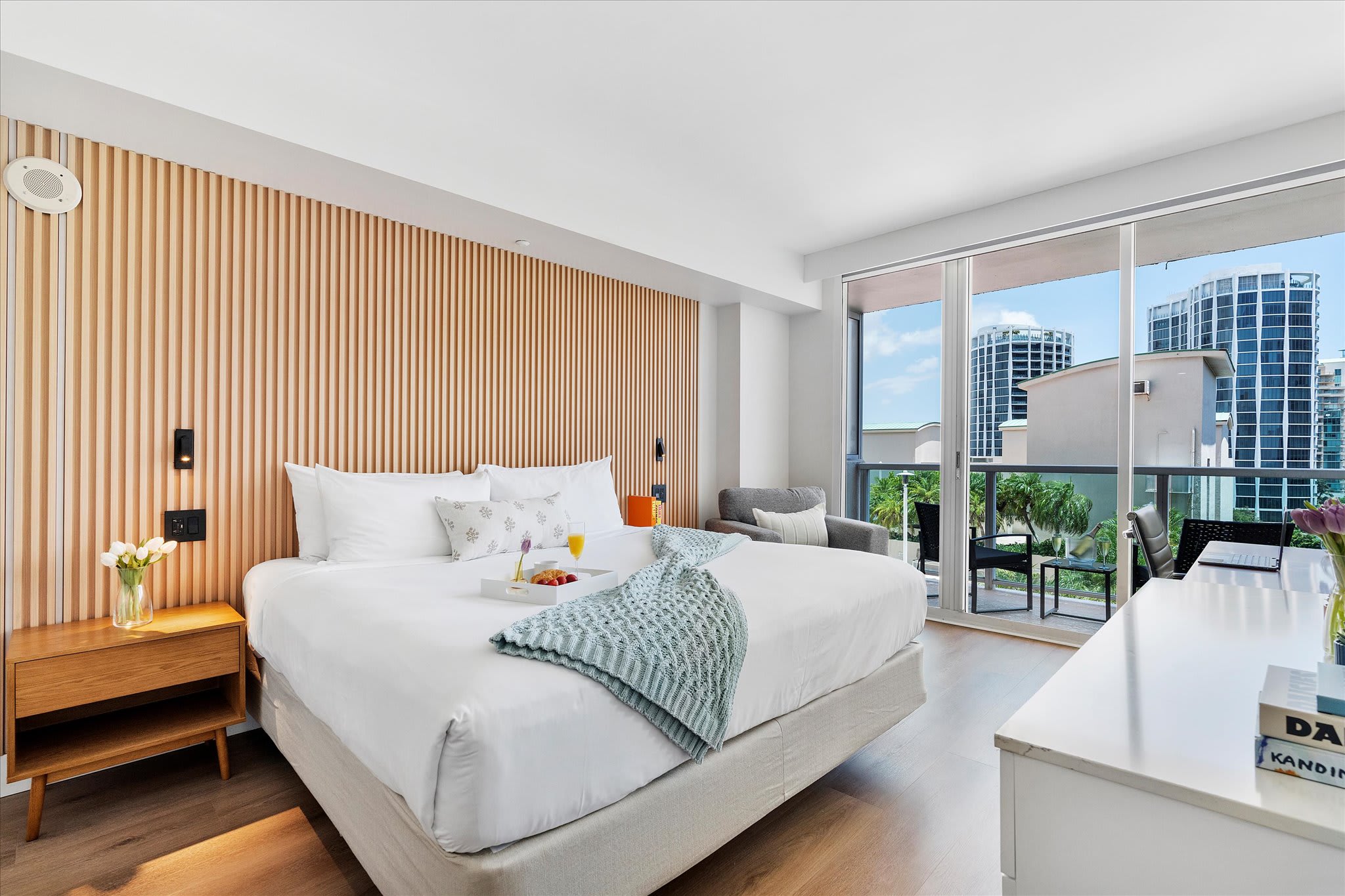 A spacious bedroom features a king-sized bed with white bedding and a light blue throw, a slatted wooden headboard accent wall, and a private balcony with city views through sliding glass doors. A breakfast tray is on the bed.