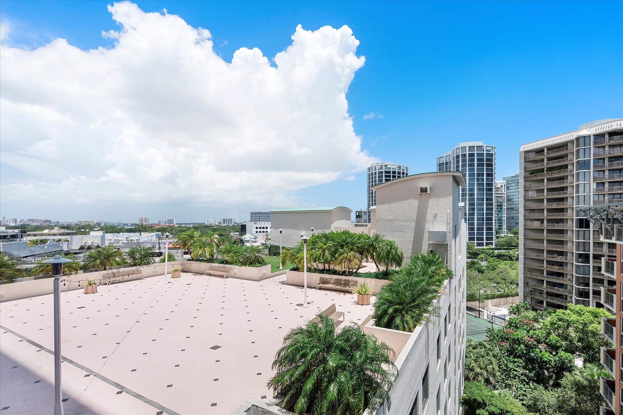 A large, open rooftop deck with light-colored paving and scattered palm trees offers views of surrounding city buildings and a bright blue sky with fluffy clouds.
