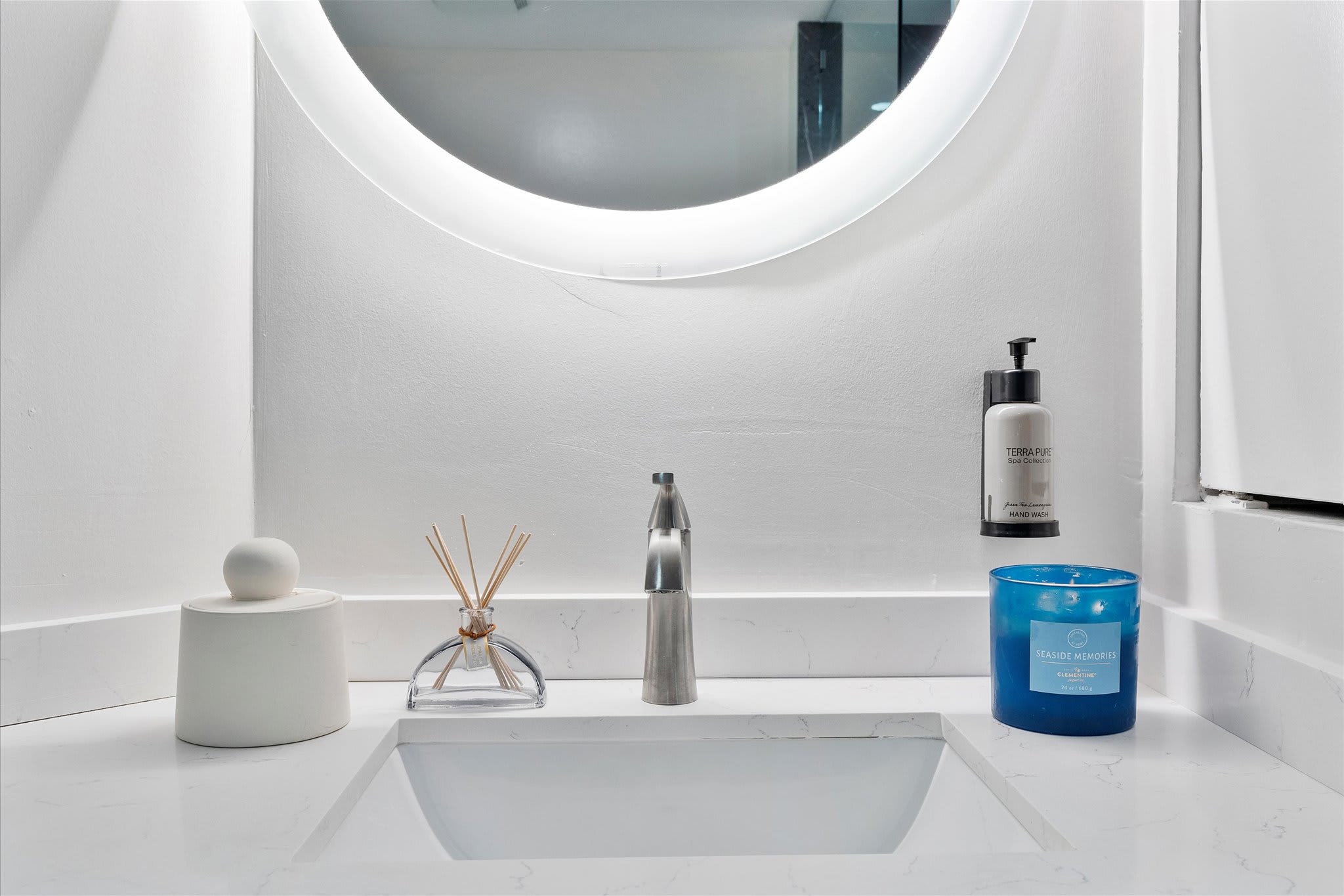 A close-up of the bathroom vanity shows a white countertop with a modern faucet, a round illuminated mirror, and decorative items including a blue candle and hand soap.