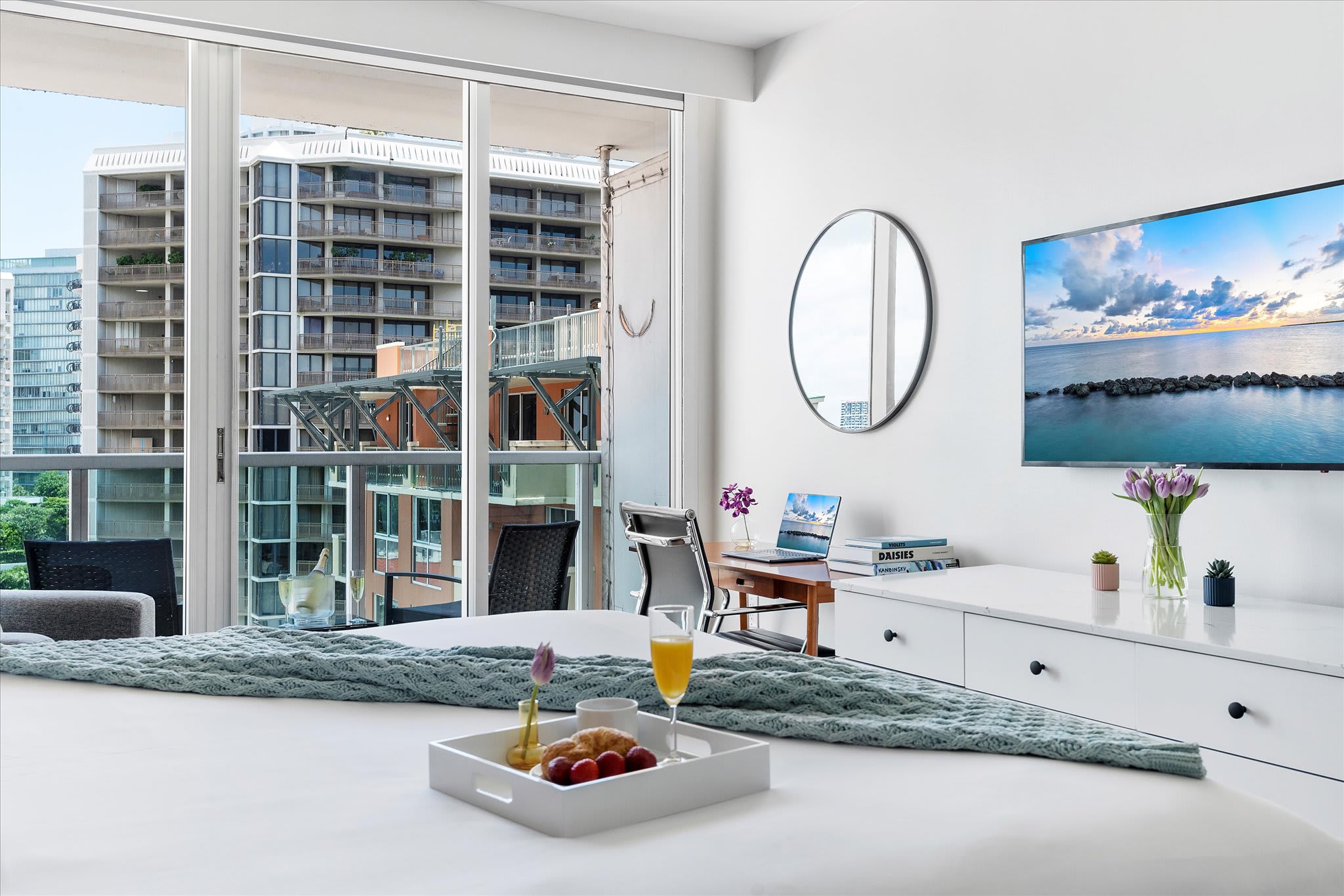 A bright bedroom with a king bed and white linens faces a large flat-screen TV mounted above a white dresser, with a wooden desk and chair to the left, and a balcony visible through sliding glass doors.
