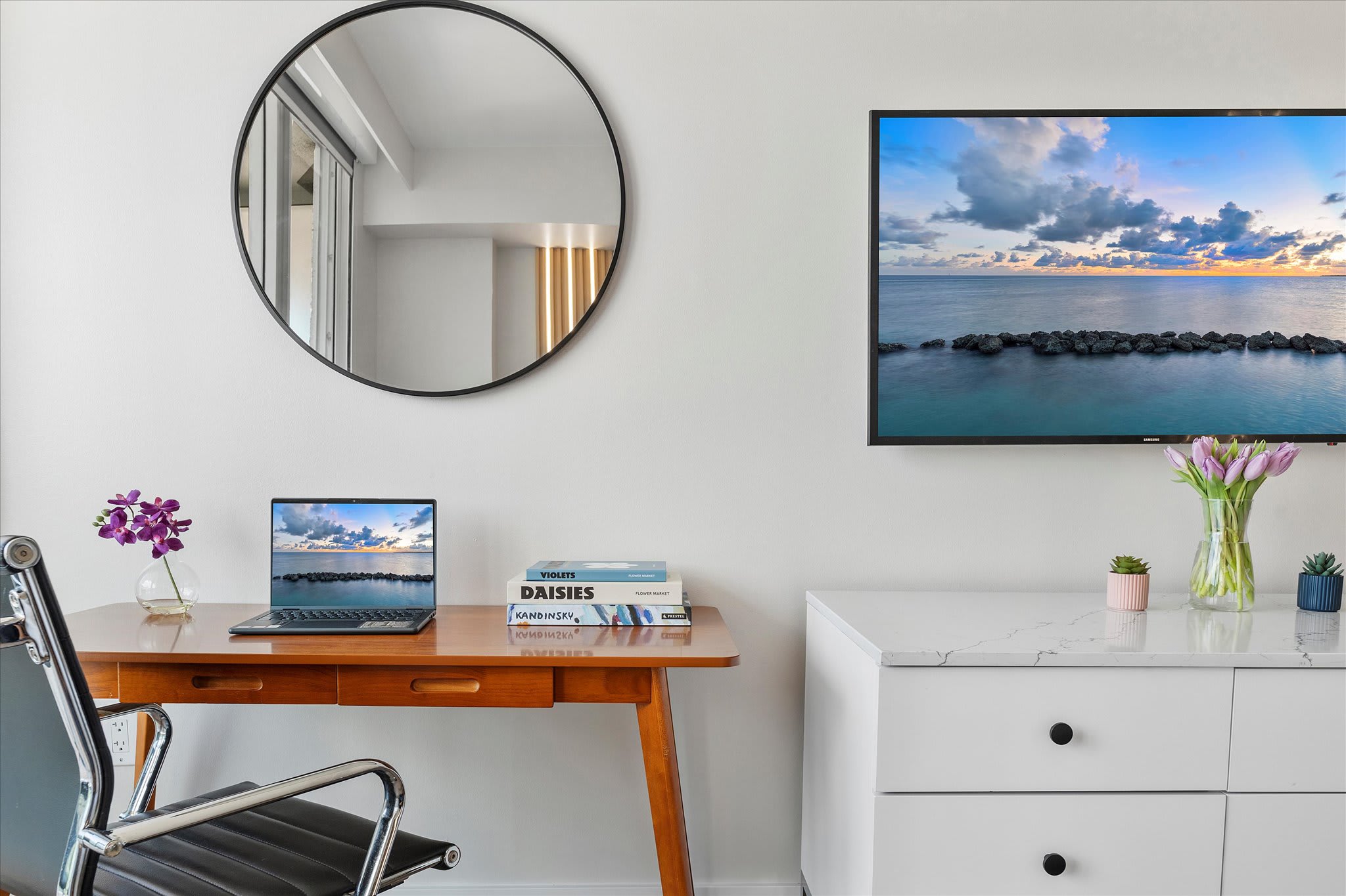 A dedicated workspace within the bedroom features a wooden desk with a laptop, books, and a vase of flowers, accompanied by a black office chair and a round mirror on the white wall. A flat-screen TV is visible to the right.