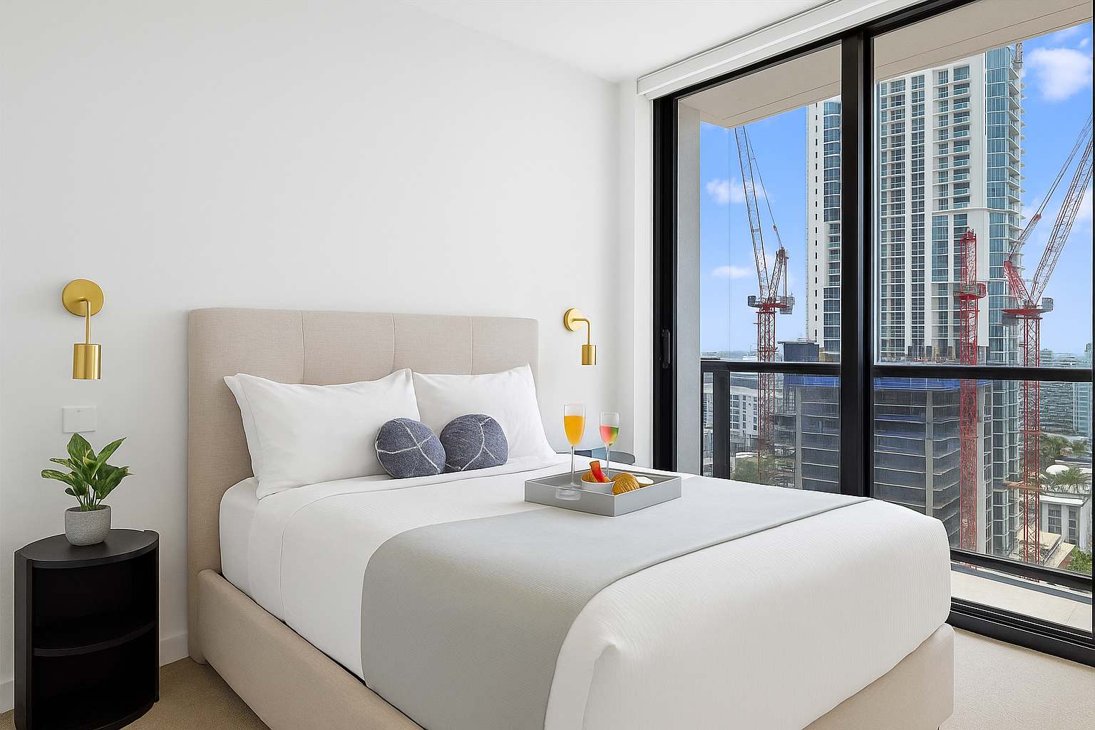 A cozy bedroom with a queen-size bed, light upholstered headboard, and modern gold wall sconces. Floor-to-ceiling windows offer a city view with construction cranes, and a breakfast tray is placed on the bed.