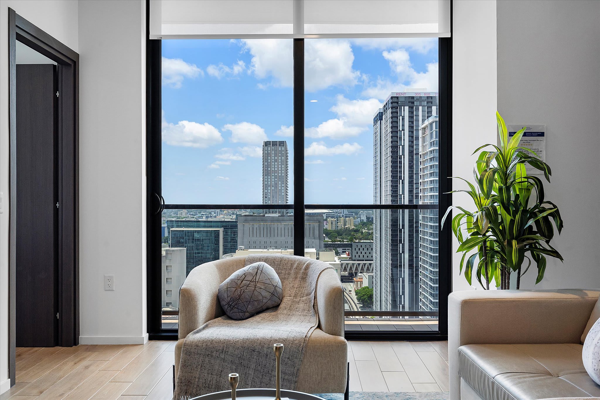 Cozy corner of the living room featuring a comfortable armchair with a throw blanket, positioned next to a large floor-to-ceiling window offering expansive views of the city skyline and surrounding buildings.