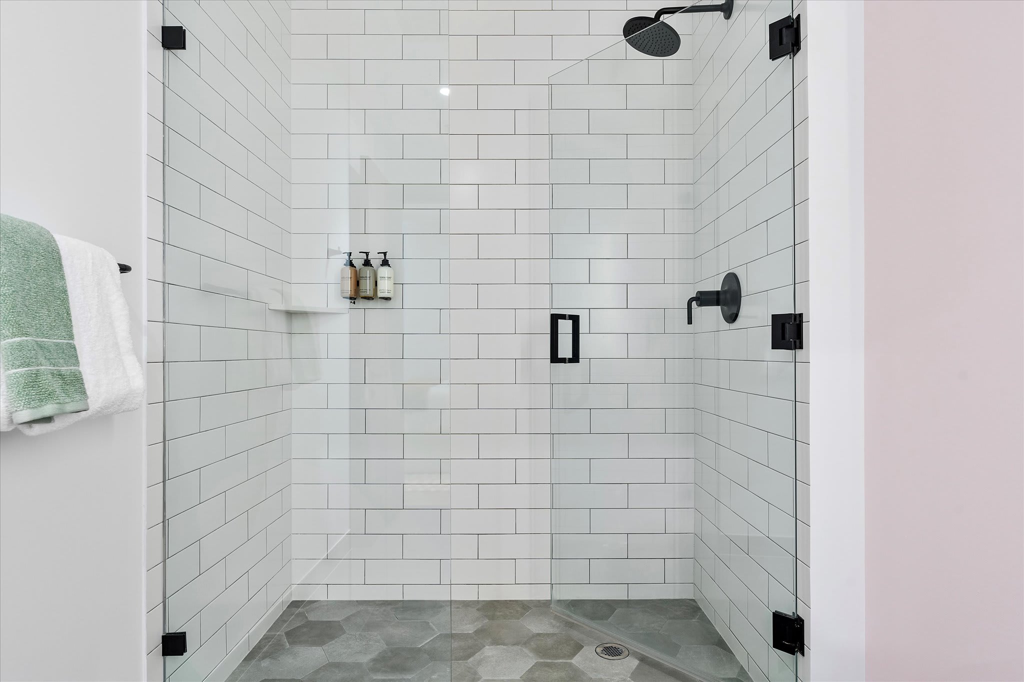 A spacious walk-in shower with white subway tiles on the walls and hexagonal gray floor tiles, enclosed by a clear glass door. Modern black fixtures include a rain showerhead and a wall-mounted shower caddy.