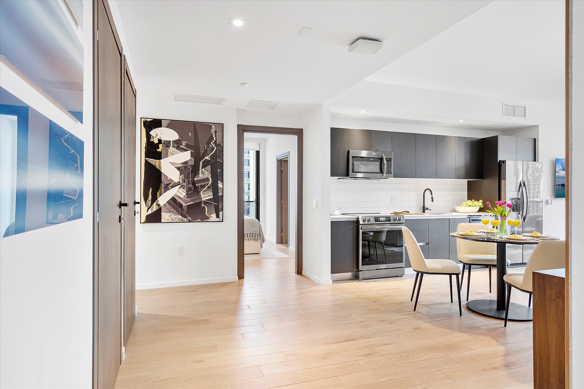 A bright open-concept space featuring light hardwood floors, a modern kitchen with dark cabinetry, and a dining area with a round table set for two. A large abstract artwork hangs on the wall, and a bedroom is visible through an open doorway.