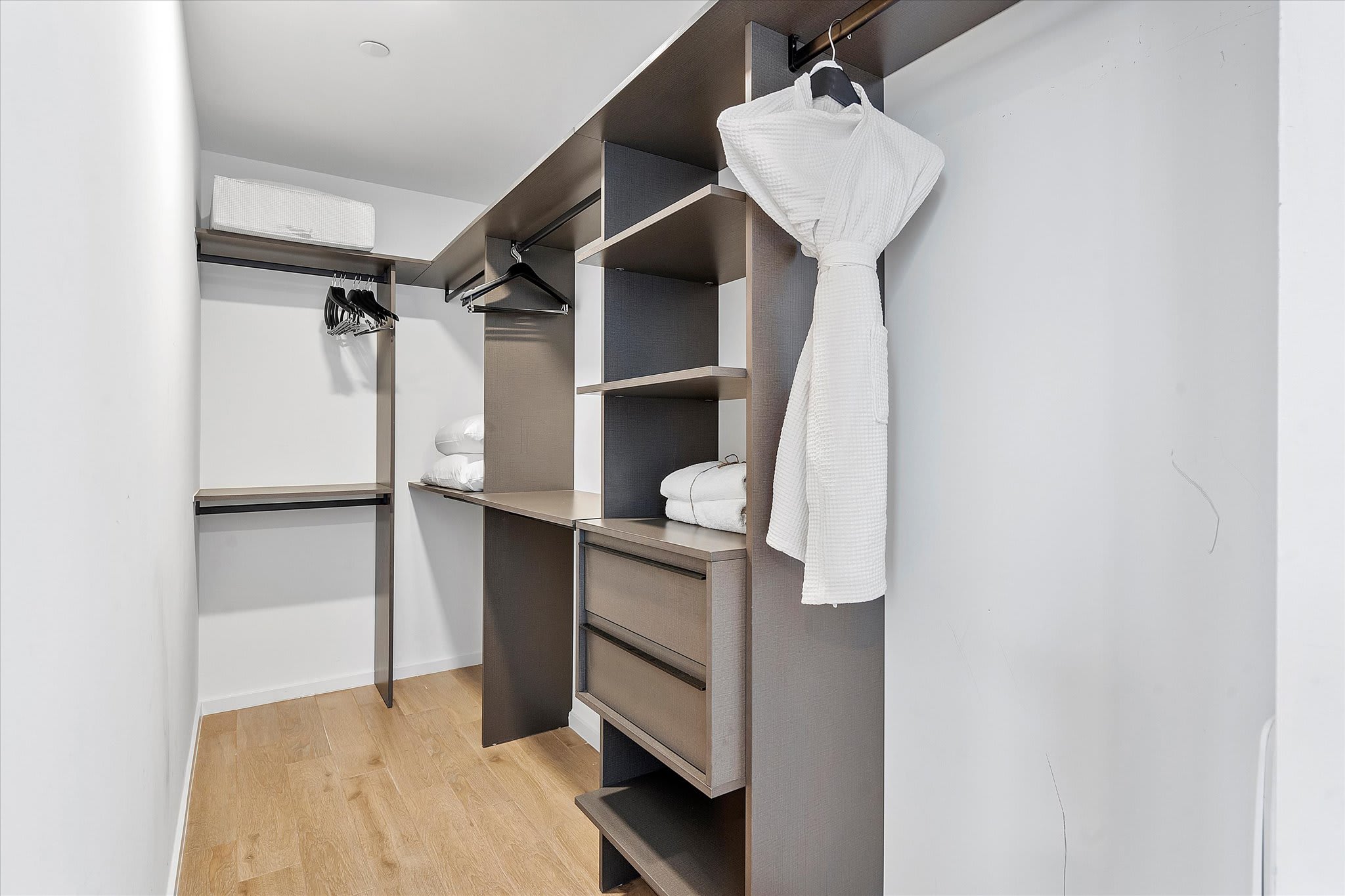 Spacious walk-in closet with dark wood shelving units, drawers, and hanging rods. A white bathrobe hangs on a hanger, and folded white towels are neatly stacked on shelves. Light wood flooring completes the organized space.