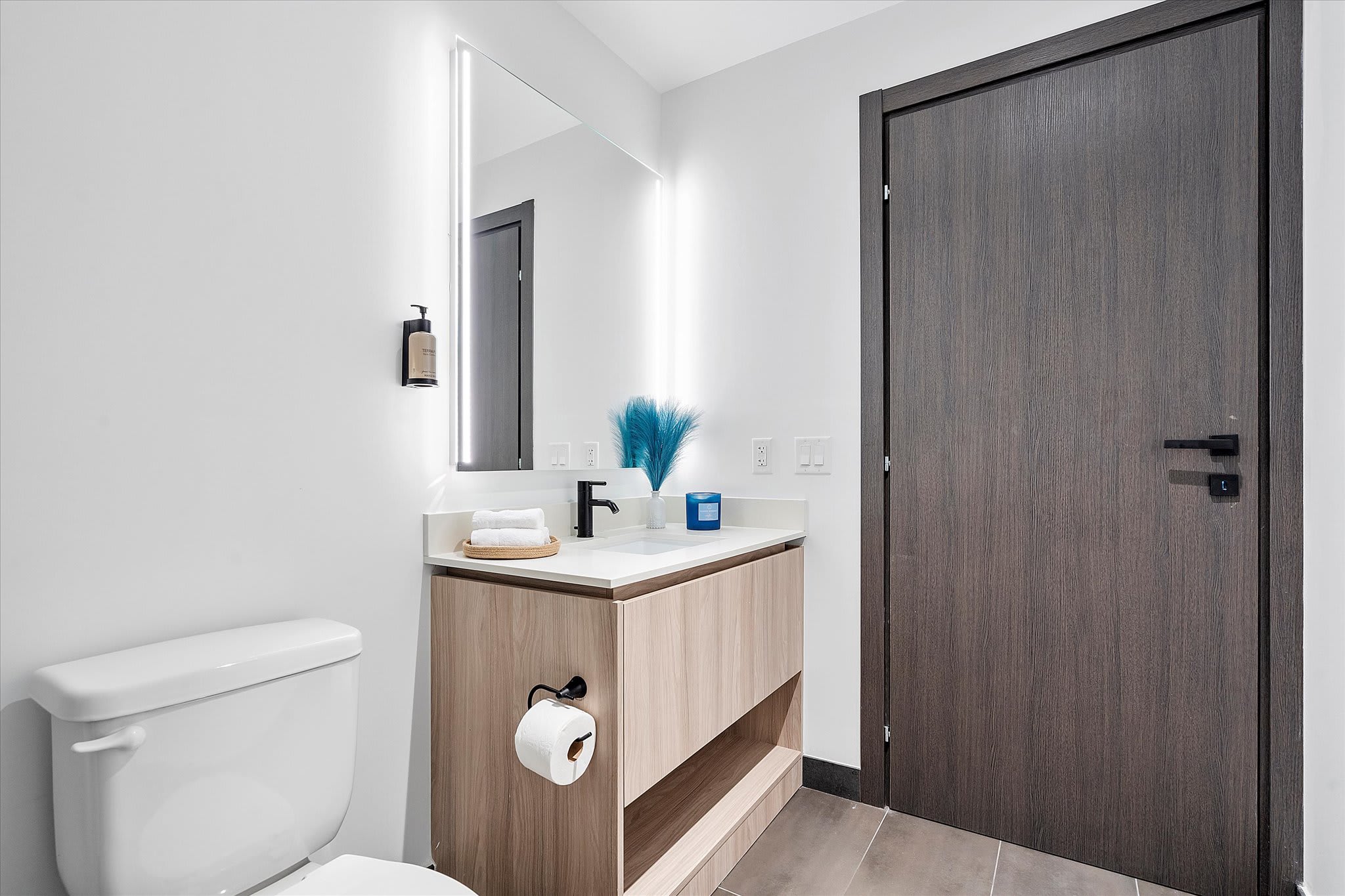 A clean, modern bathroom featuring a light wood vanity with a white countertop, a large illuminated mirror, and a toilet. Dark gray floor tiles and a dark wood door complete the contemporary look.