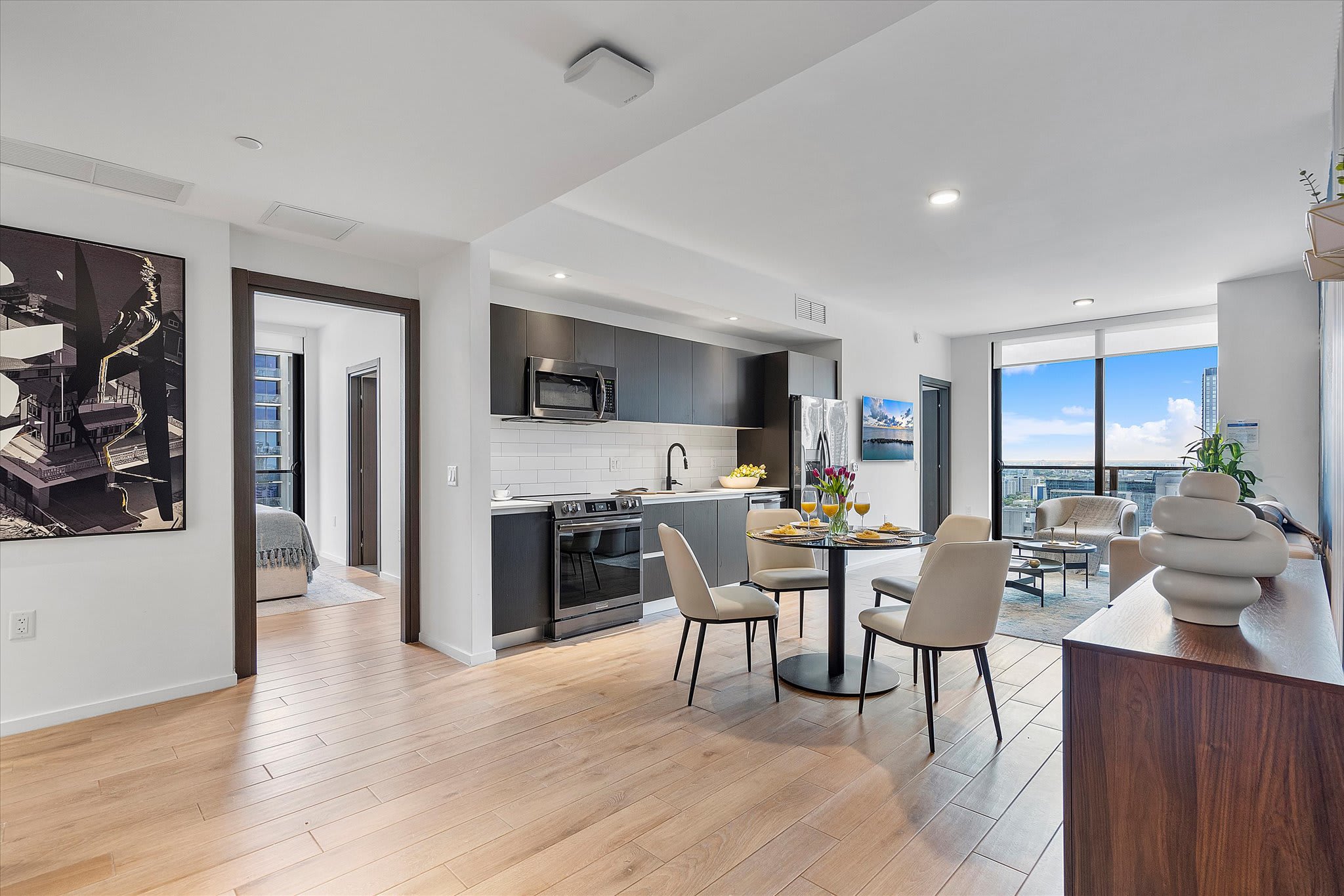 Open-concept living space combining a modern kitchen with dark cabinetry and stainless steel appliances, a dining area with a round table and four chairs, and a bright living room with large windows offering city views.