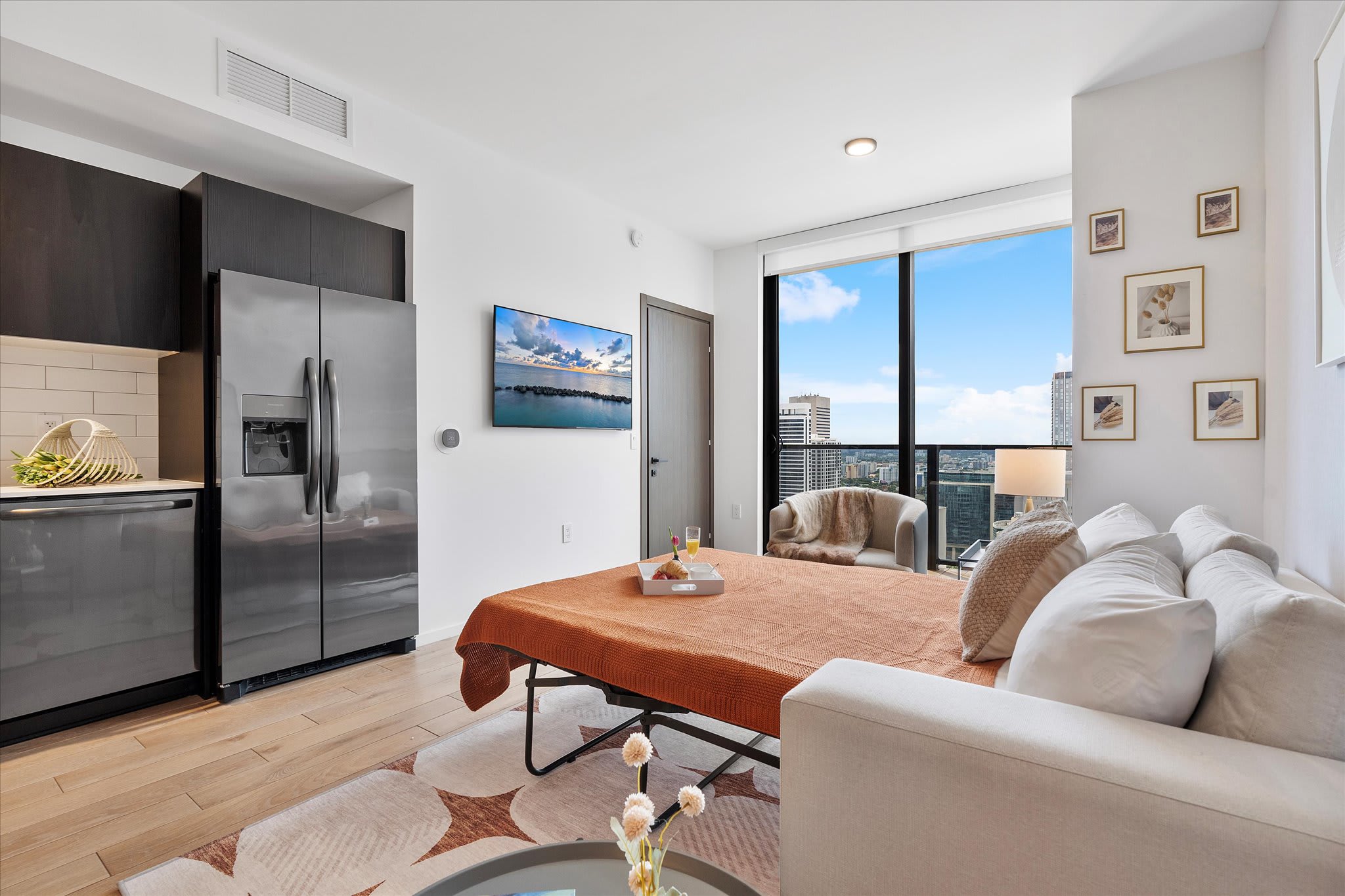Versatile living space featuring a sofa and a pull-out bed, with a wall-mounted TV and a stainless steel refrigerator visible. Large windows provide a bright atmosphere and city views, indicating a flexible sleeping arrangement.