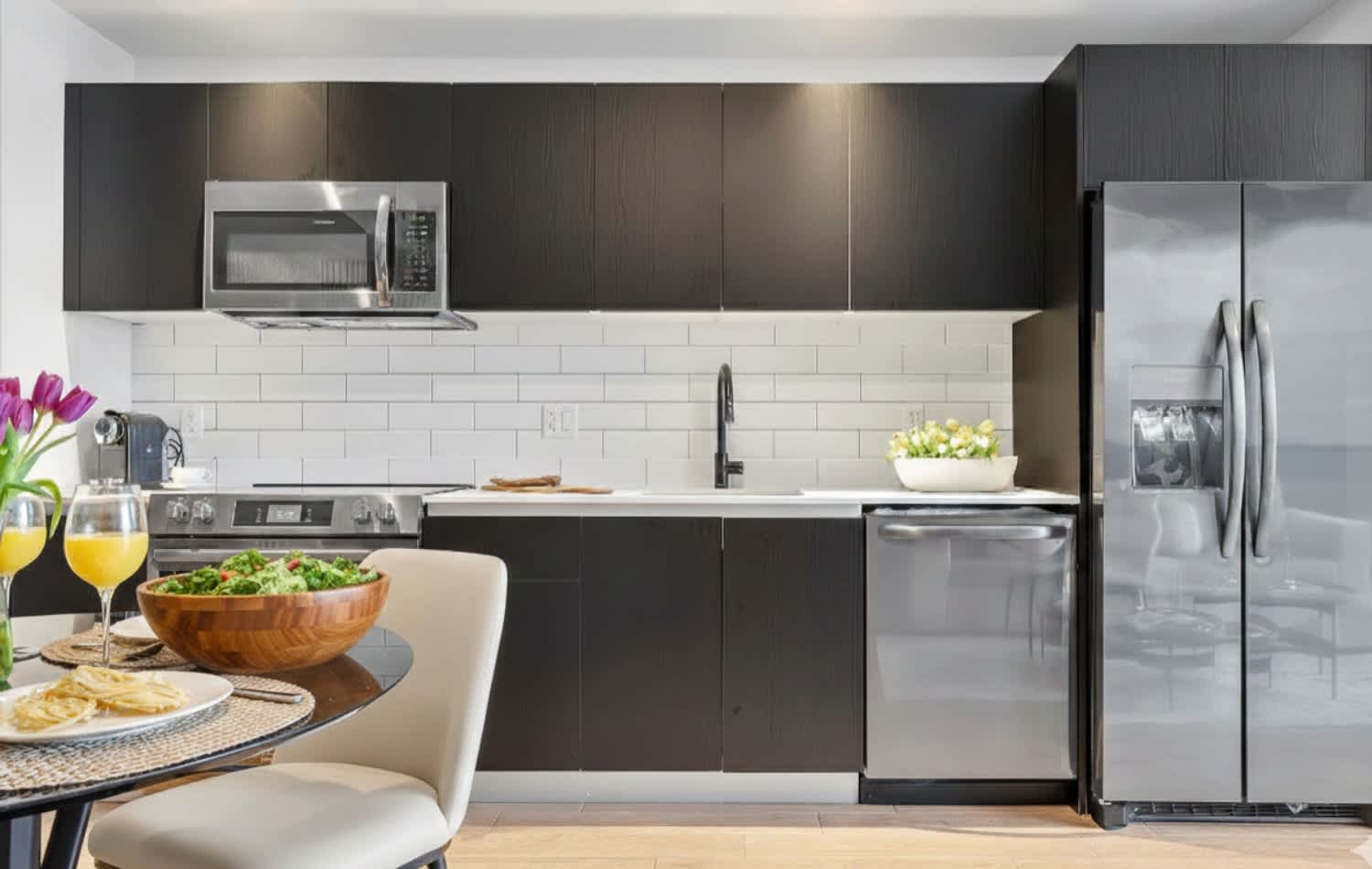 Contemporary kitchen with dark flat-panel cabinetry, white subway tile backsplash, and stainless steel appliances including a microwave, range, dishwasher, and a large refrigerator. A small dining table with chairs is visible in the foreground.