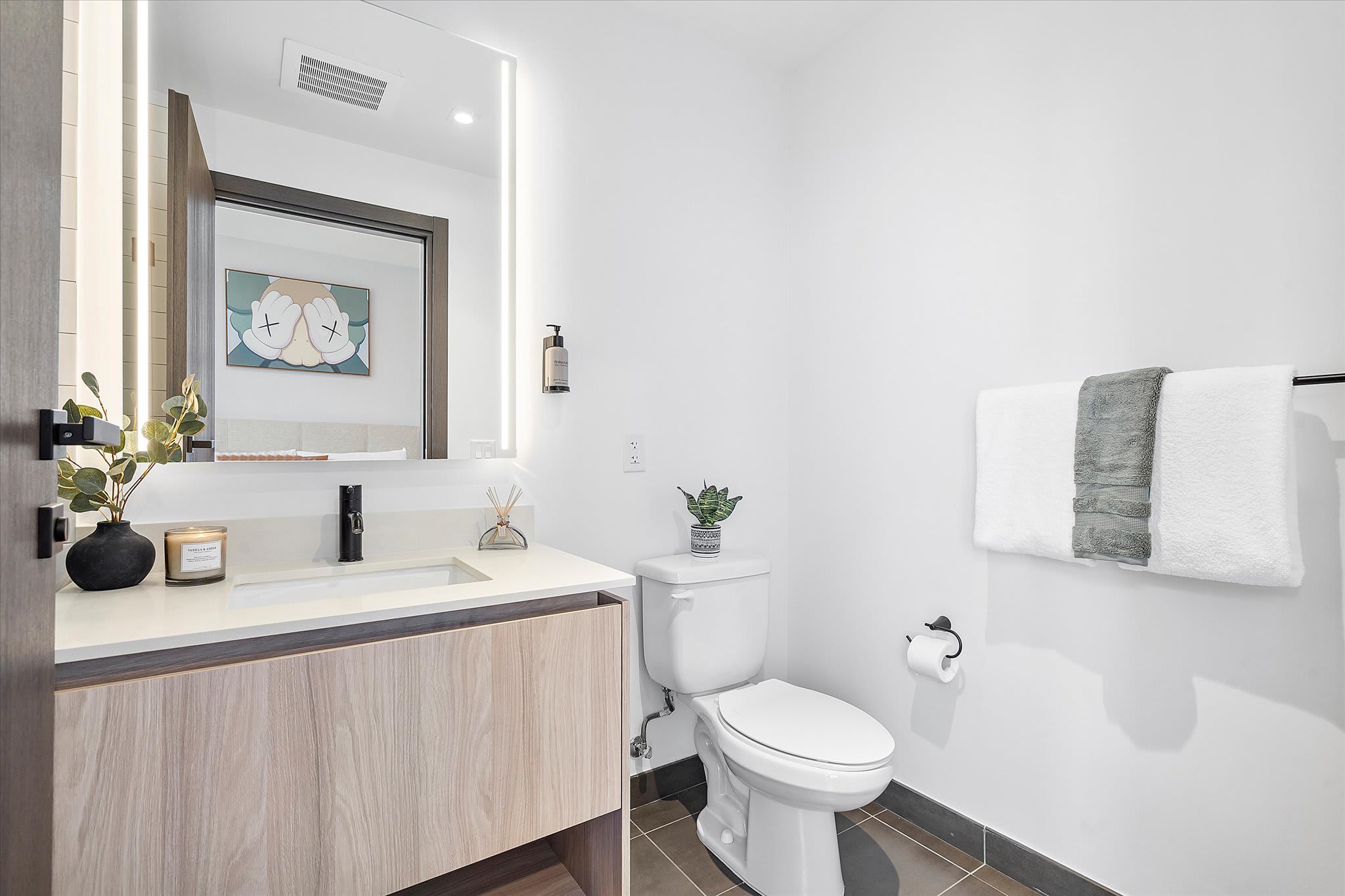 Modern bathroom featuring a light wood vanity with a white countertop and sink, an illuminated rectangular mirror, a white toilet, and dark gray floor tiles. A small plant and toiletries adorn the vanity, and white and gray towels hang on a wall rack.