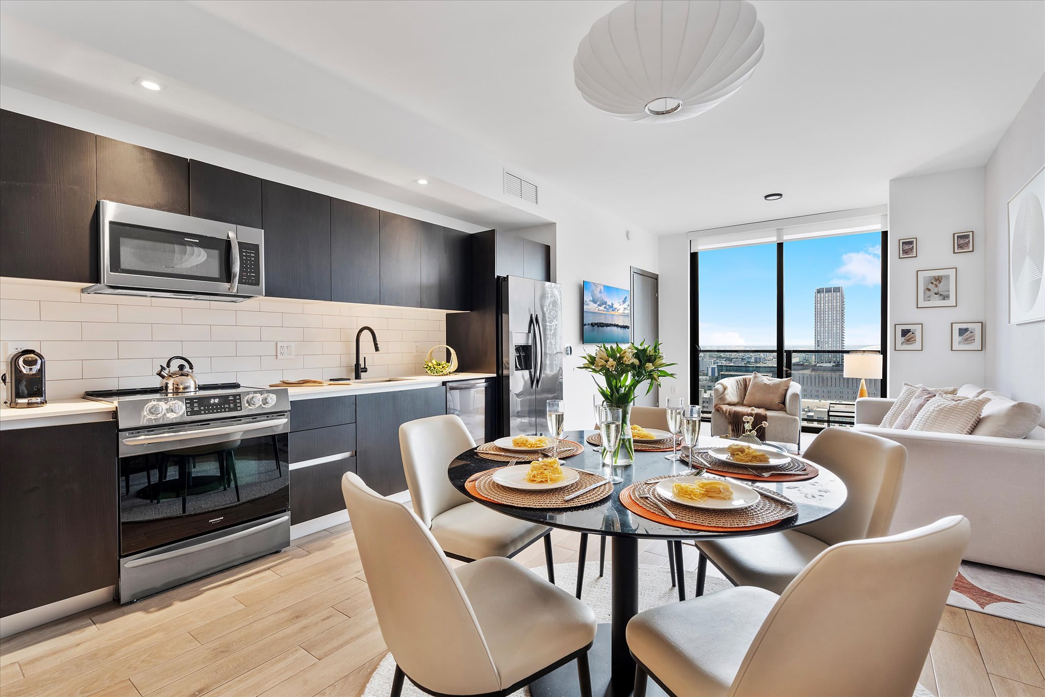 Elegant dining area featuring a round black dining table set for four, with light-colored chairs, adjacent to a modern kitchen with dark cabinetry and stainless steel appliances. Large windows in the background offer city views.