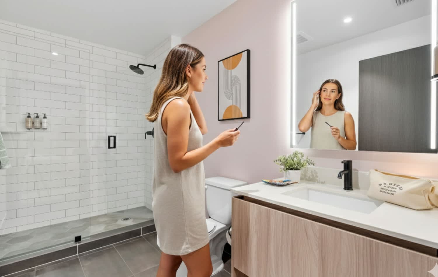 Modern bathroom with a light pink accent wall, a large illuminated mirror above a vanity with a white countertop, and a spacious walk-in shower with white subway tiles and a glass enclosure.