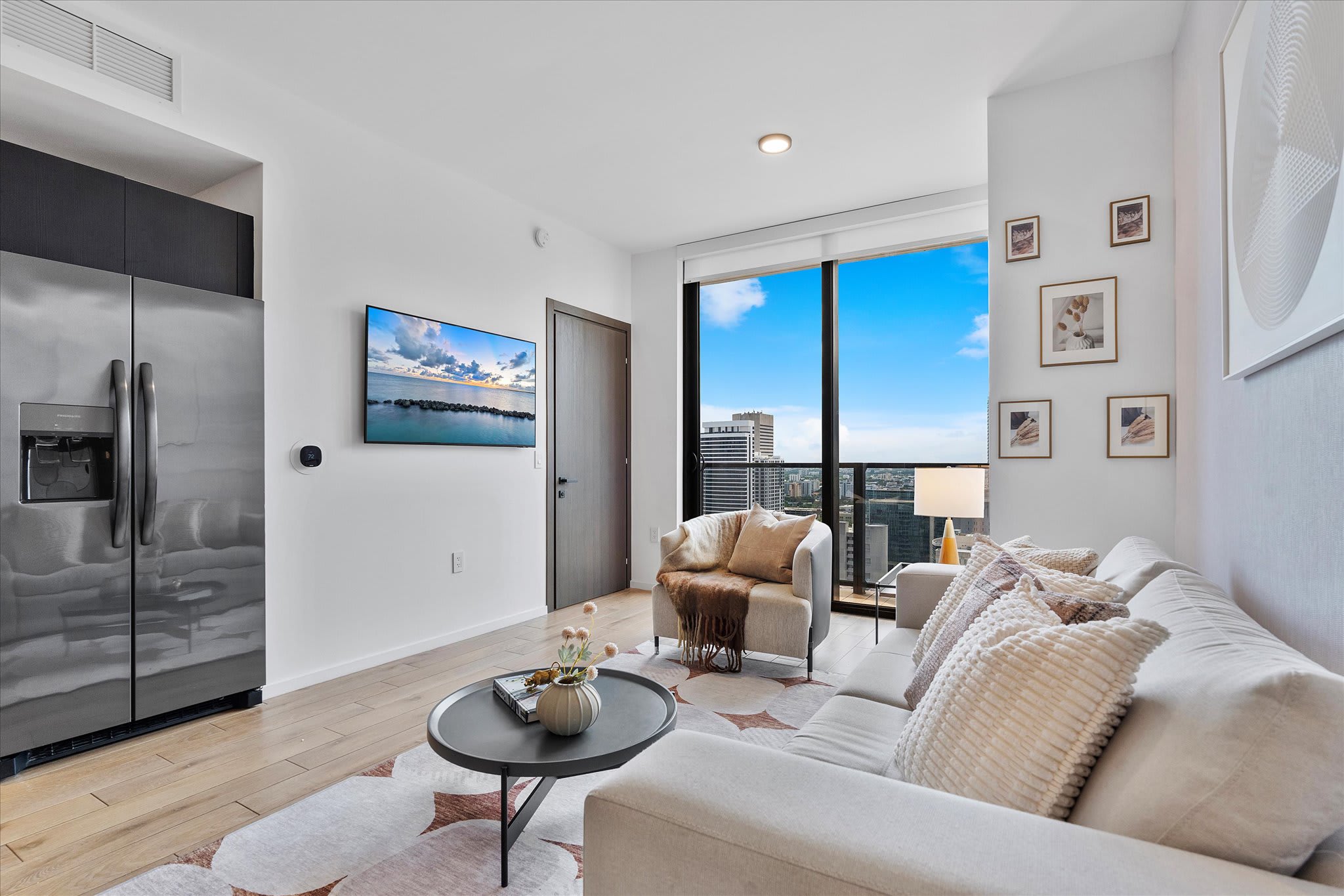 Bright and modern living room with light wood flooring, a large L-shaped sofa, an armchair, and a wall-mounted TV. A stainless steel refrigerator is visible on the left, and large windows offer a city view.